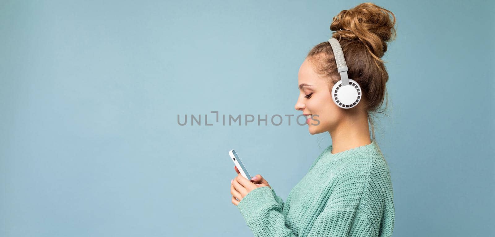 Panoramic Side-profile photo shot of beautiful joyful smiling young female person wearing stylish casual outfit isolated over colorful background wall wearing white bluetooth wireless earphones and listening to music and using mobile phone looking at gadjet display. copy space