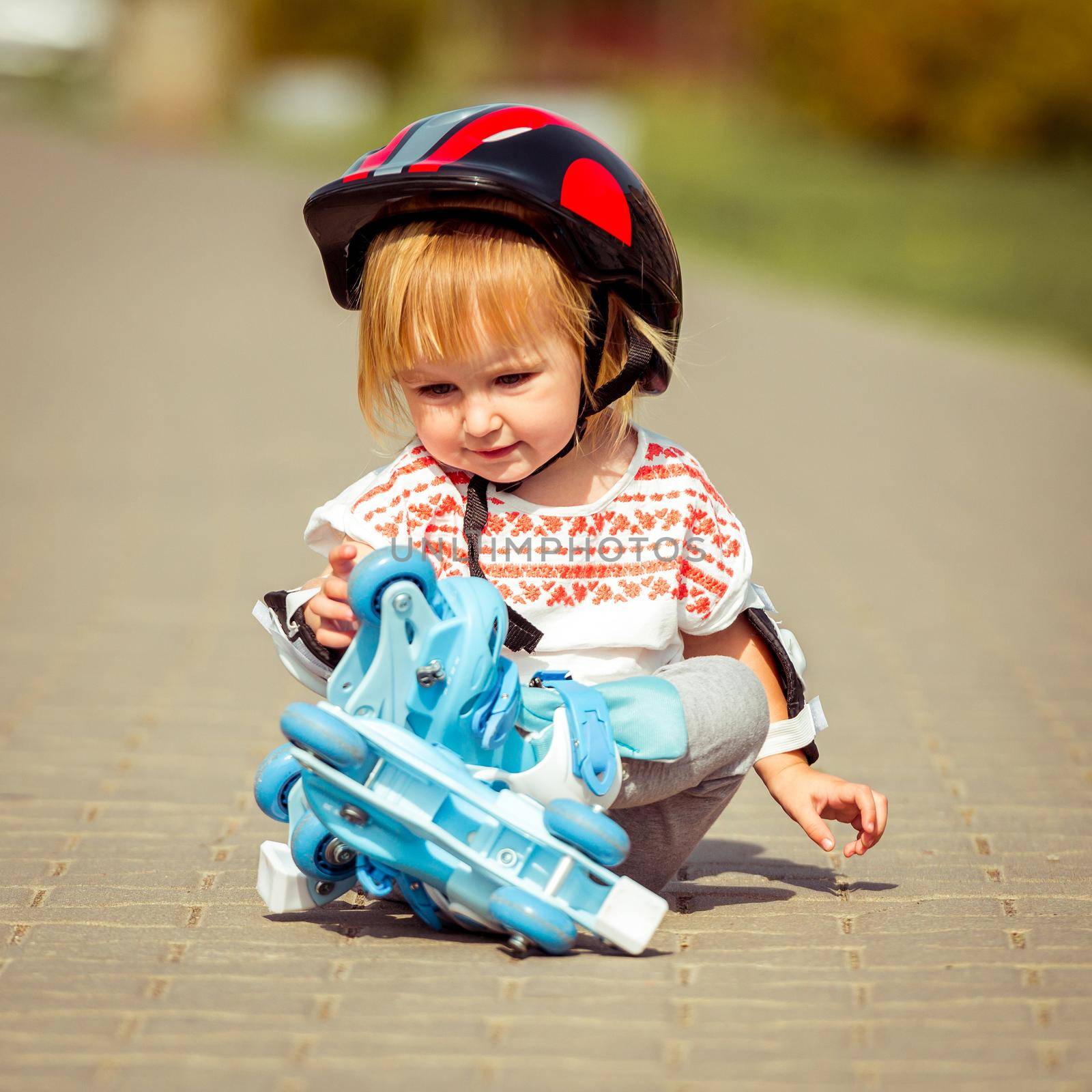 two year old pretty girl in roller skates and a helmet sitting on the street