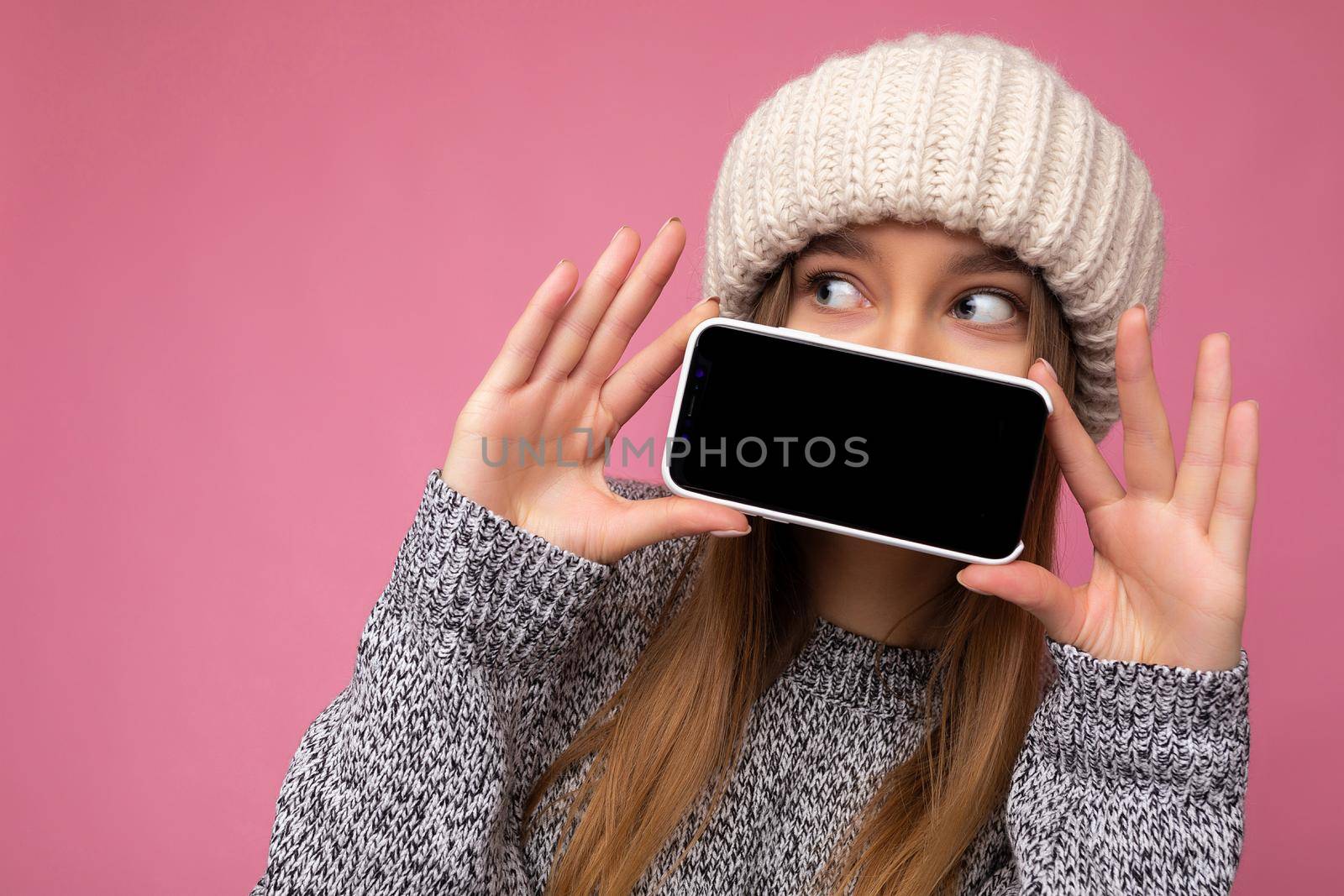 Closeup photo of Beautiful young blonde woman wearing casual grey sweater and beige hat isolated over pink background holding in hand and showing mobile phone with empty display for mockup looking to the side.