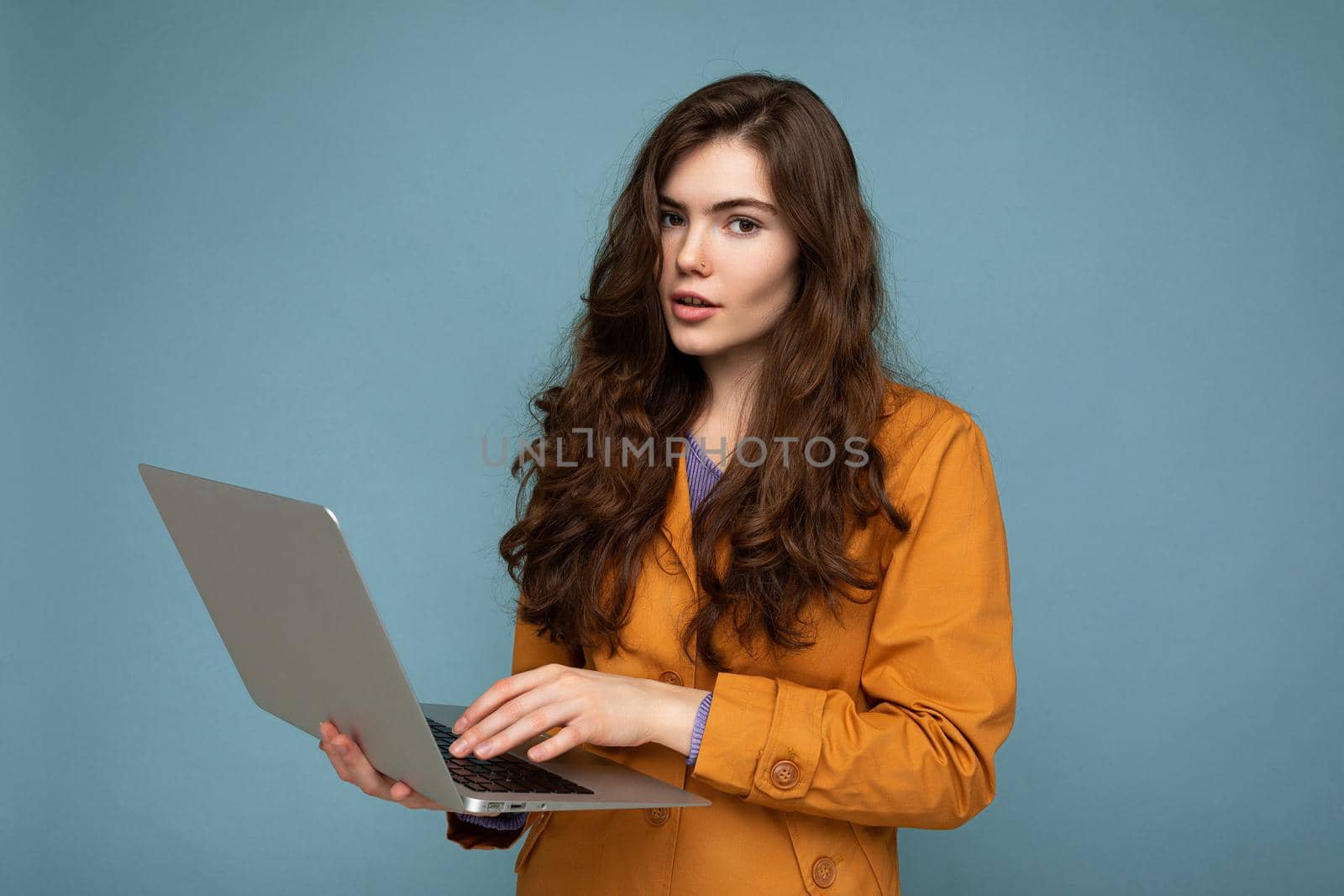 Close-up portrait of Beautiful calm brunet curly young woman holding netbook computer looking at camera wearing yellow jacket typing on keyboard isolated on blue background.