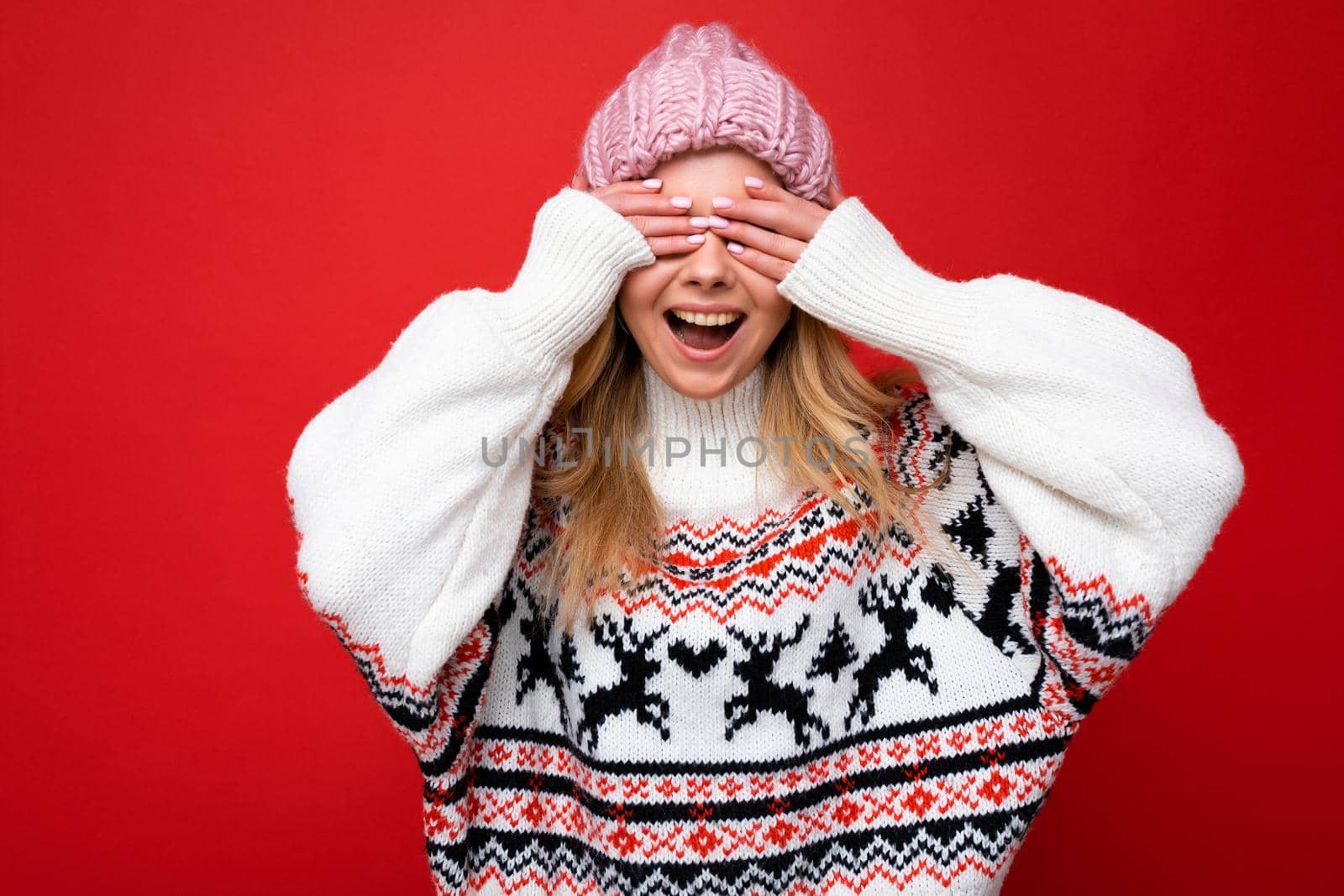 Photo of young positive happy beautiful blonde lady with sincere emotions wearing pink knitted hat and winter pullover isolated over red background with empty space and covering eyes with hands.