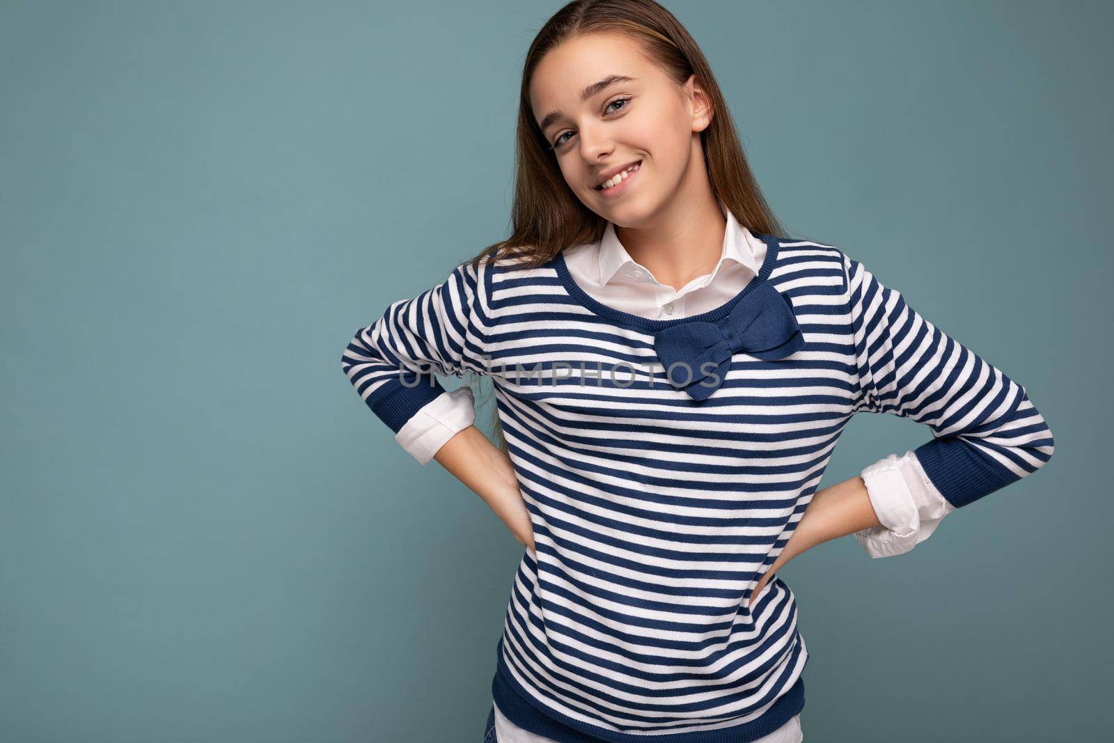 Shot of beautiful positive smiling brunette little girl wearing striped longsleeve standing isolated over blue background wall and looking at camera. Empty space, copy space