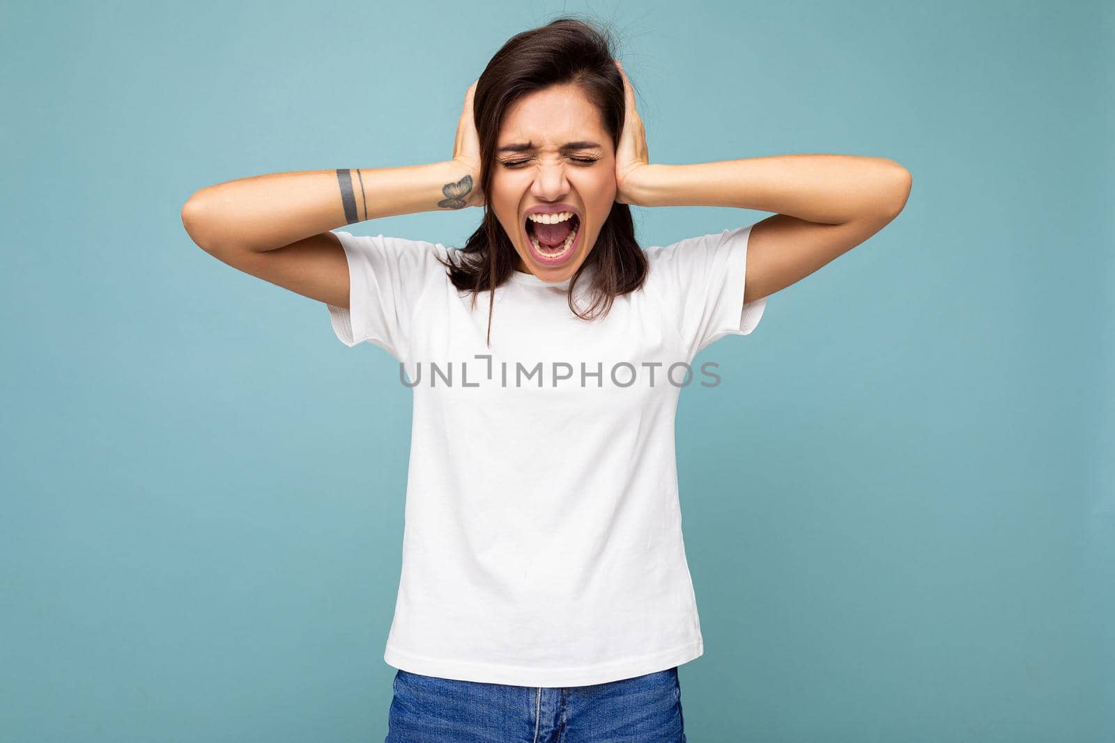 Portrait of emotional young beautiful brunette woman with sincere emotions wearing casual white t-shirt for mockup isolated on blue background with empty space and covering ears with hands and shouting.