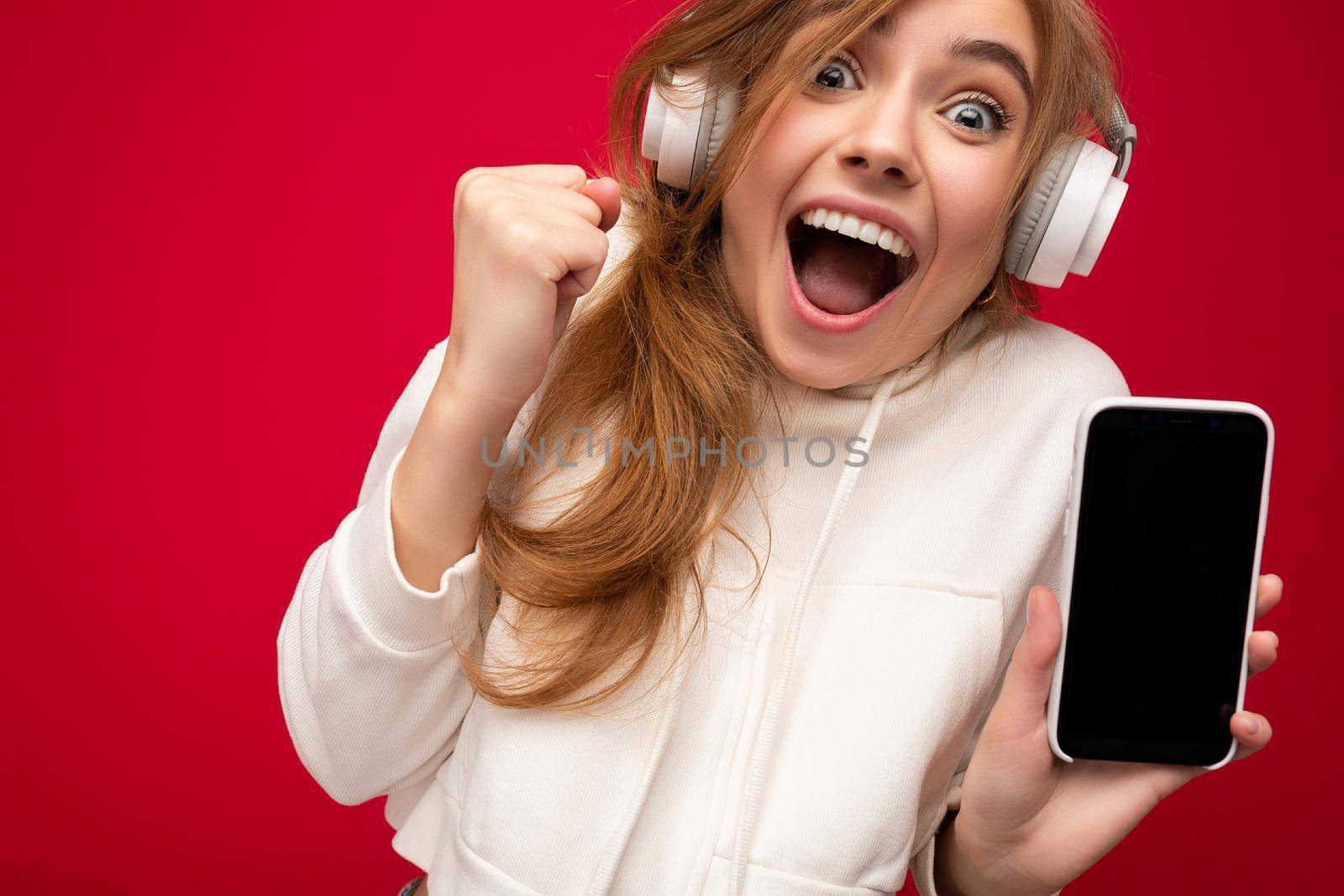 Closeup portrait of shocked emotional attractive young blond woman wearing white hoodie isolated over colorful background wall holding and showing mobile phone with empty screen for cutout and wearing bluetooth headphones listening to pop music looking at camera and shouting.