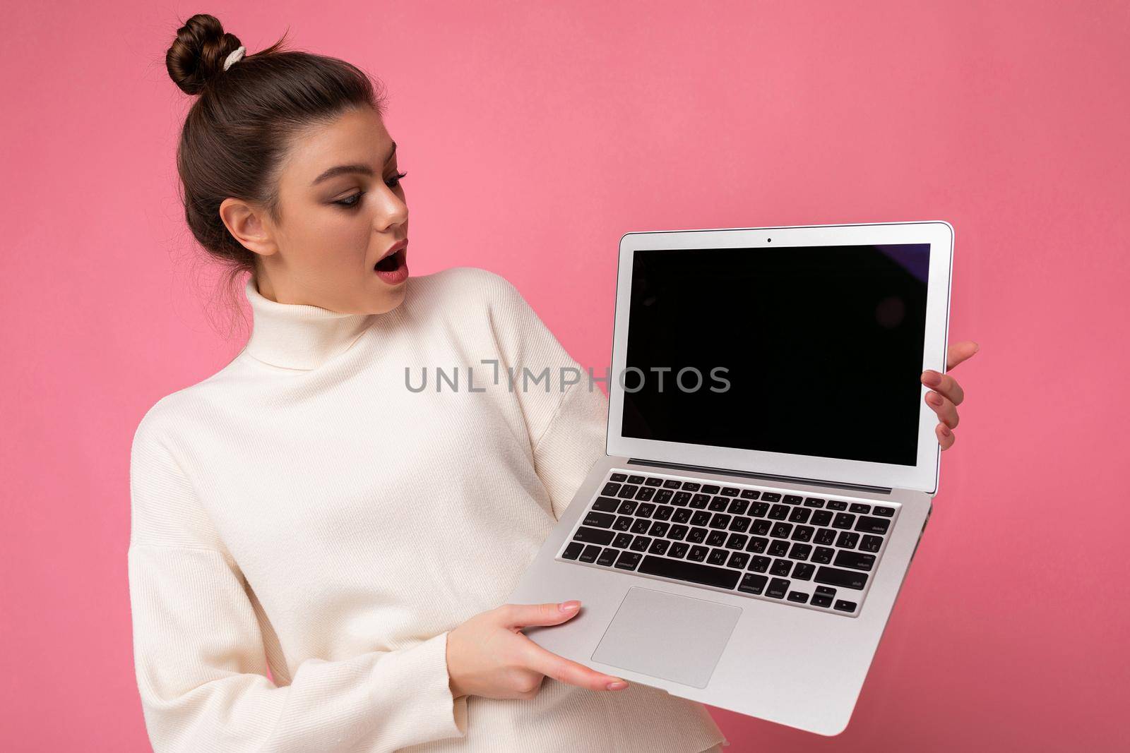 Photo of beautiful amazed and surprised woman with gathered brunette hair wearing white sweater holding computer laptop and looking at the open netbook isolated over pink wall background.