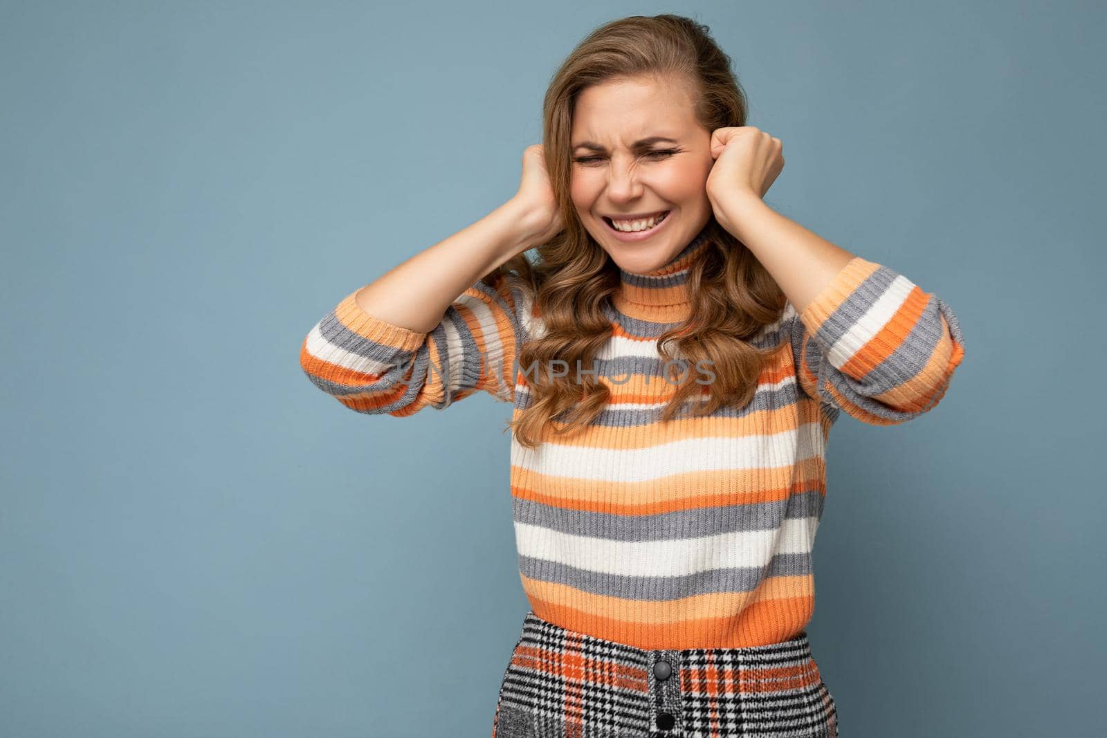 Photo of emotional young attractive blonde curly woman with sincere emotions wearing casual striped pullover isolated on blue background with copy space and covering ears trying not to hear by TRMK