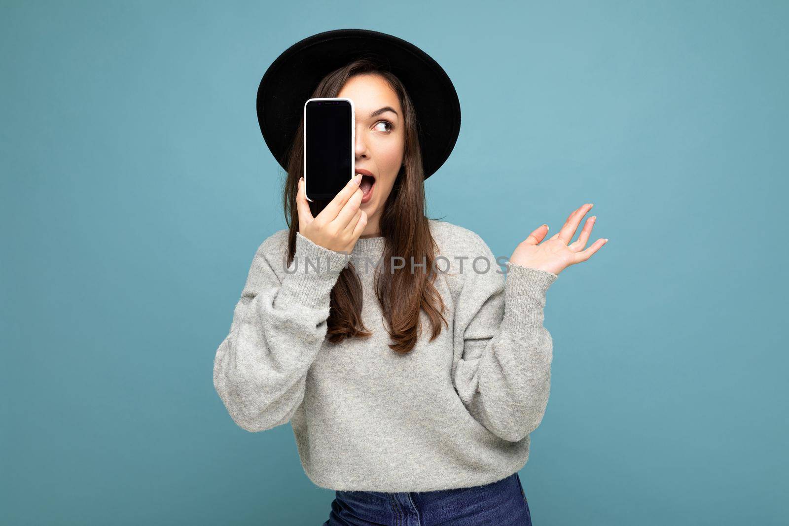 Amazed Beautiful positive woman wearing black hat and grey sweater holding mobilephone showing smartphone isolated on background looking to the side with open mouth.Mock up, cutout, free space. copy space