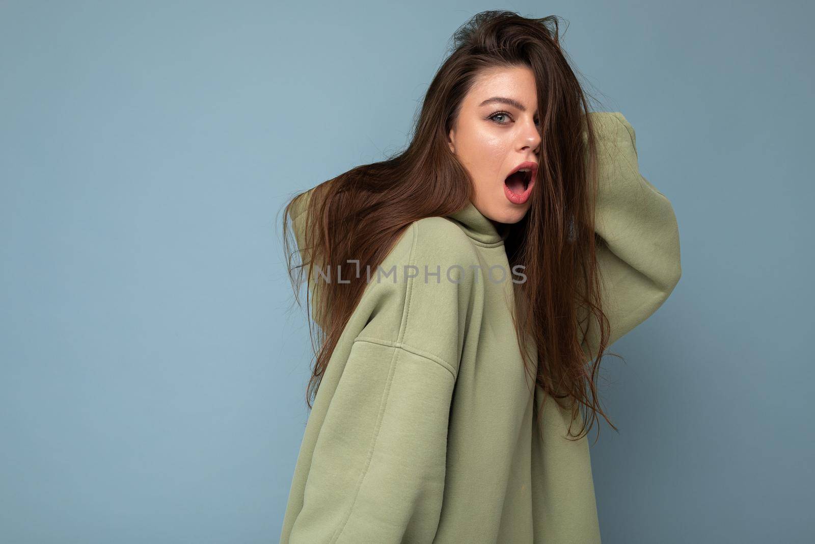 Young shocked amazed beautiful woman looking at camera. Trendy girl in casual hipster hoodie clothes. Positive female shows facial emotions. Funny model isolated on blue background with free space for text by TRMK