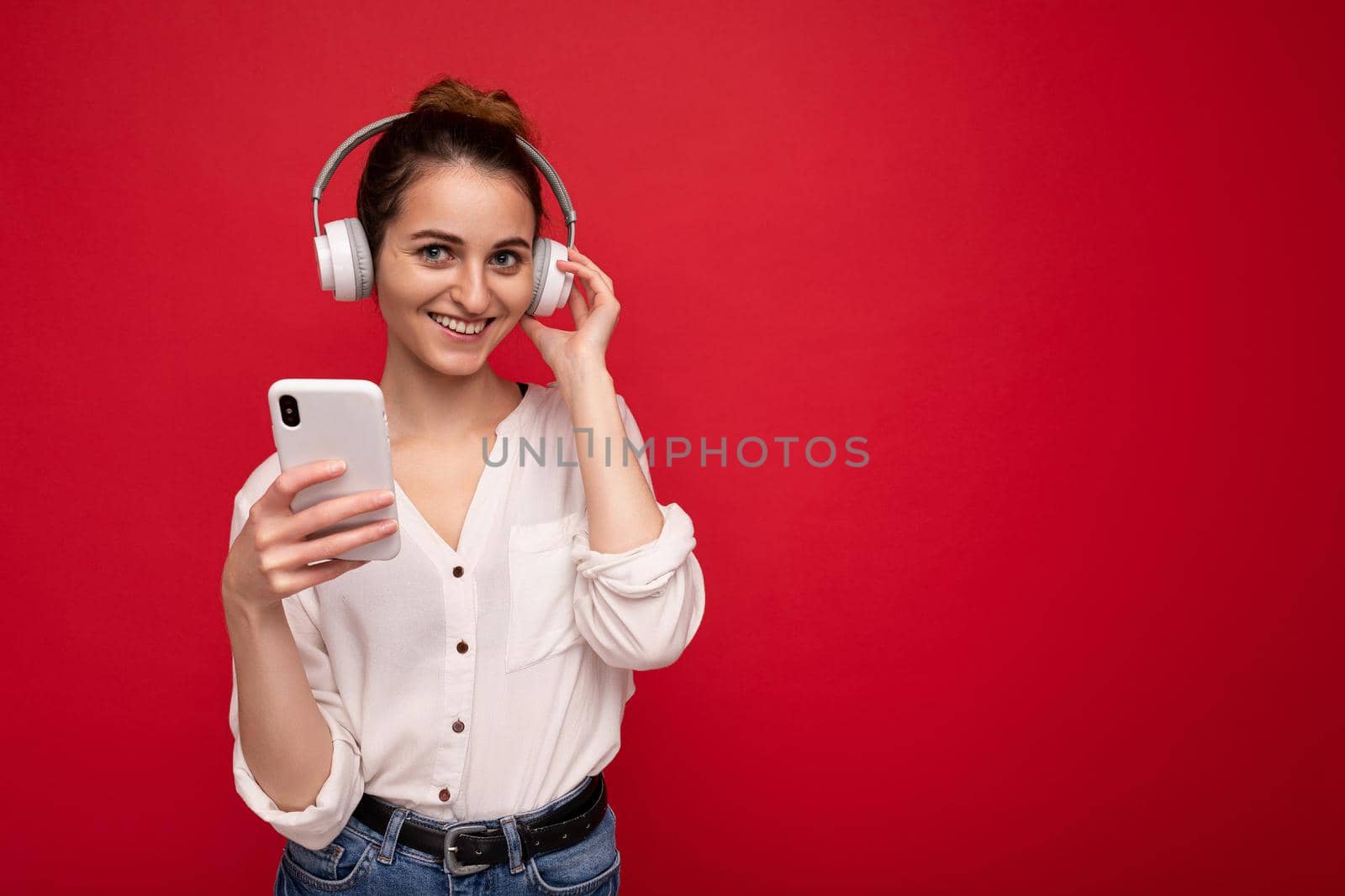 Photo shot of beautiful joyful smiling young female person wearing stylish casual outfit isolated over colorful background wall wearing white bluetooth wireless earphones and listening to music and using mobile phone looking at camera. copy space