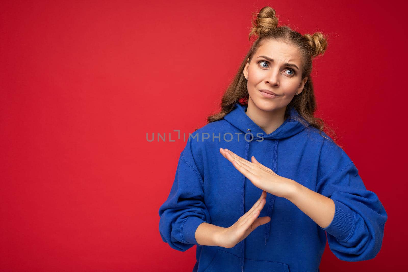 Photo of emotional adult attractive blonde woman with two horns with sincere emotions wearing hipster bright blue hoodie isolated over red background with copy space and showing timeout sign by TRMK