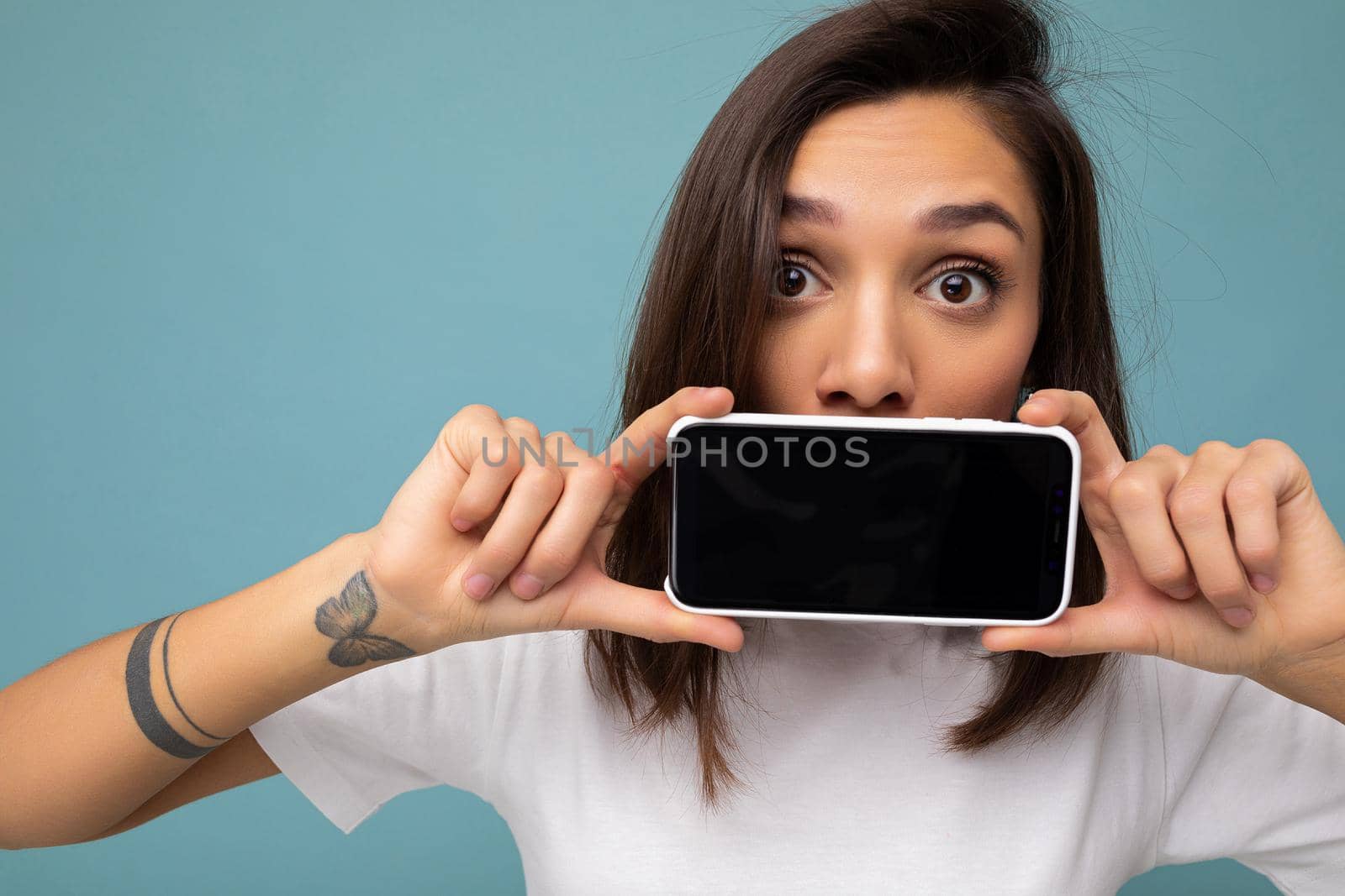 Photo of pretty surprised young brunette woman good looking wearing white t-shirt standing isolated on blue background with copy space holding smartphone showing phone in hand with empty screen screen for cutout looking at camera.