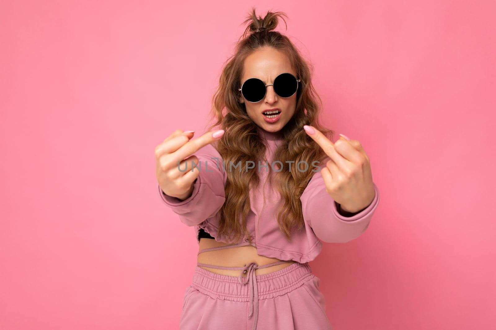 Young self-confident cool beautiful blonde wavy-haired woman with sincere emotions wearing stylish pink sport suit and sunglasses isolated on pink background with copy space and showing middle finger.