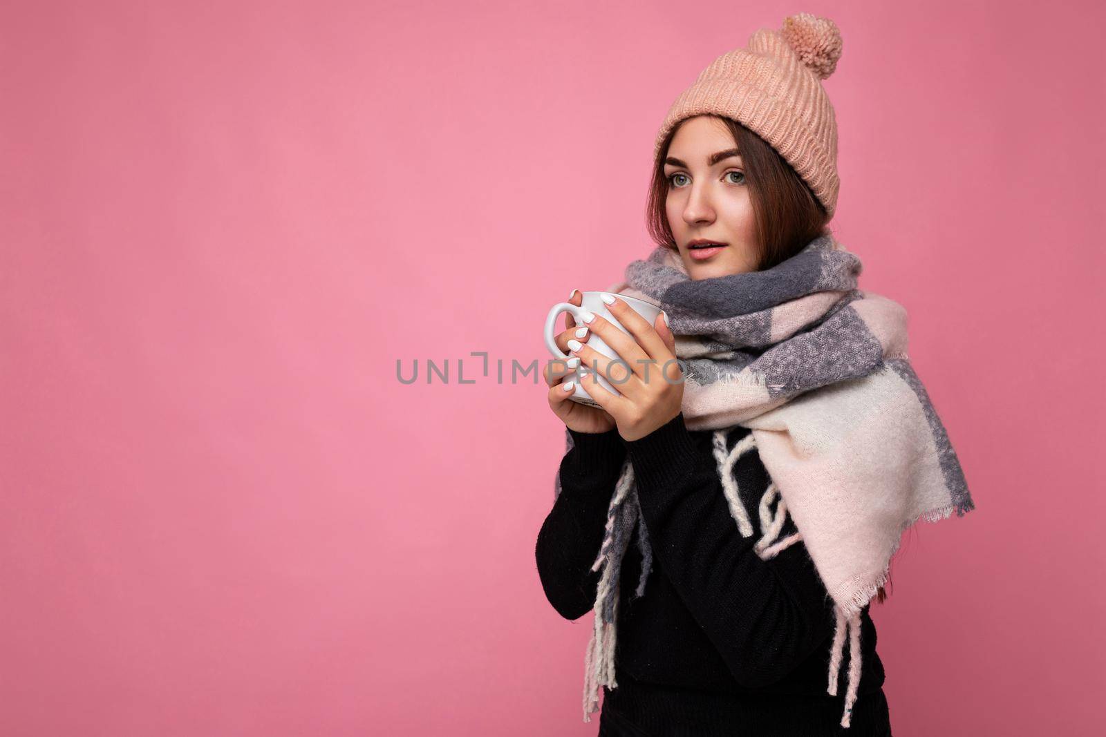 Beautiful young brunet woman wearing black sweater hat and warm scarf isolated over pink background holding paper mug for mockup drinking hot tea and looking at camera and having cold. empty space