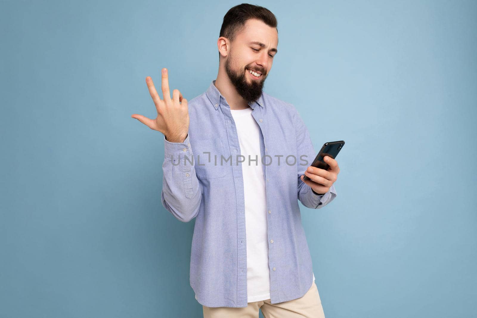 Photo shot of handsome happy smiled satisfued positive good looking young man wearing casual stylish outfit poising isolated on background with empty space holding in hand and using mobile phone messaging sms looking at smartphone display screen by TRMK
