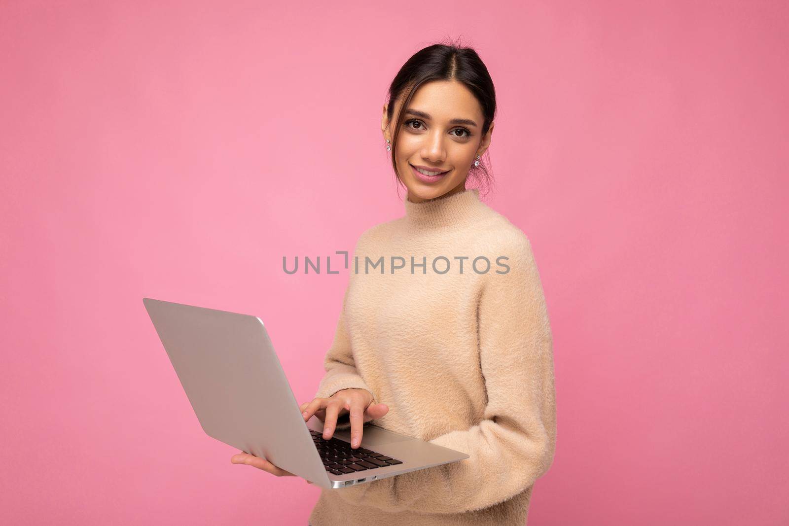 Photo of Beautiful young brunette woman wearing beige sweater holding netbook computer typing text on keyboard looking at camera isolated over pink background by TRMK