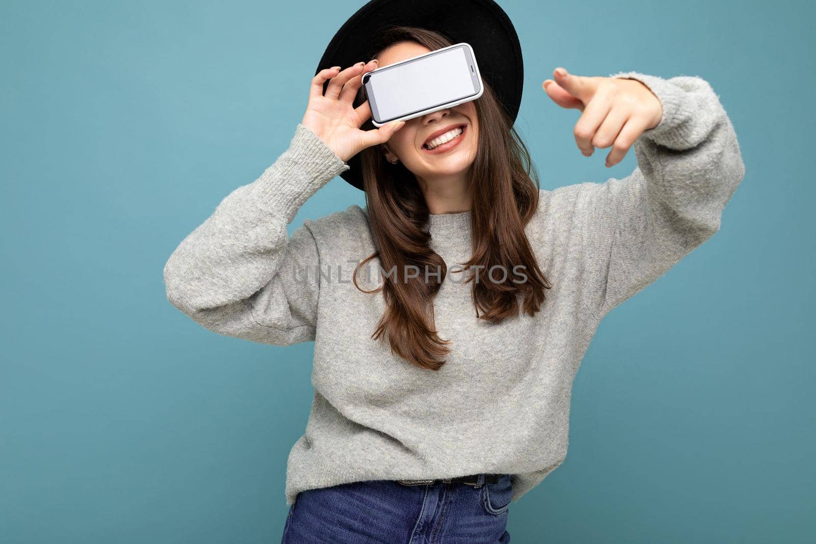 Closeup of attractive young happy woman wearing black hat and grey sweater having fun holding phone pointing finger finger at camera isolated on background.Mock up, cutout, empty space