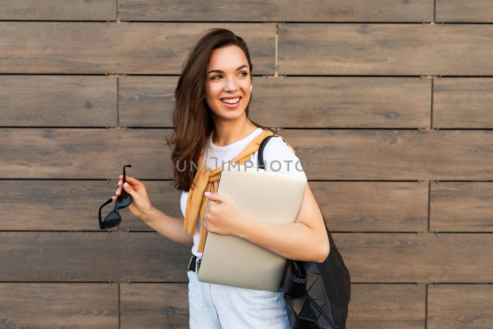 Close-up portrait of nice-looking charming attractive lovely fascinating cheerful cheery straight-haired brunet woman looking to side in the street with laptop computer and glasses in hands