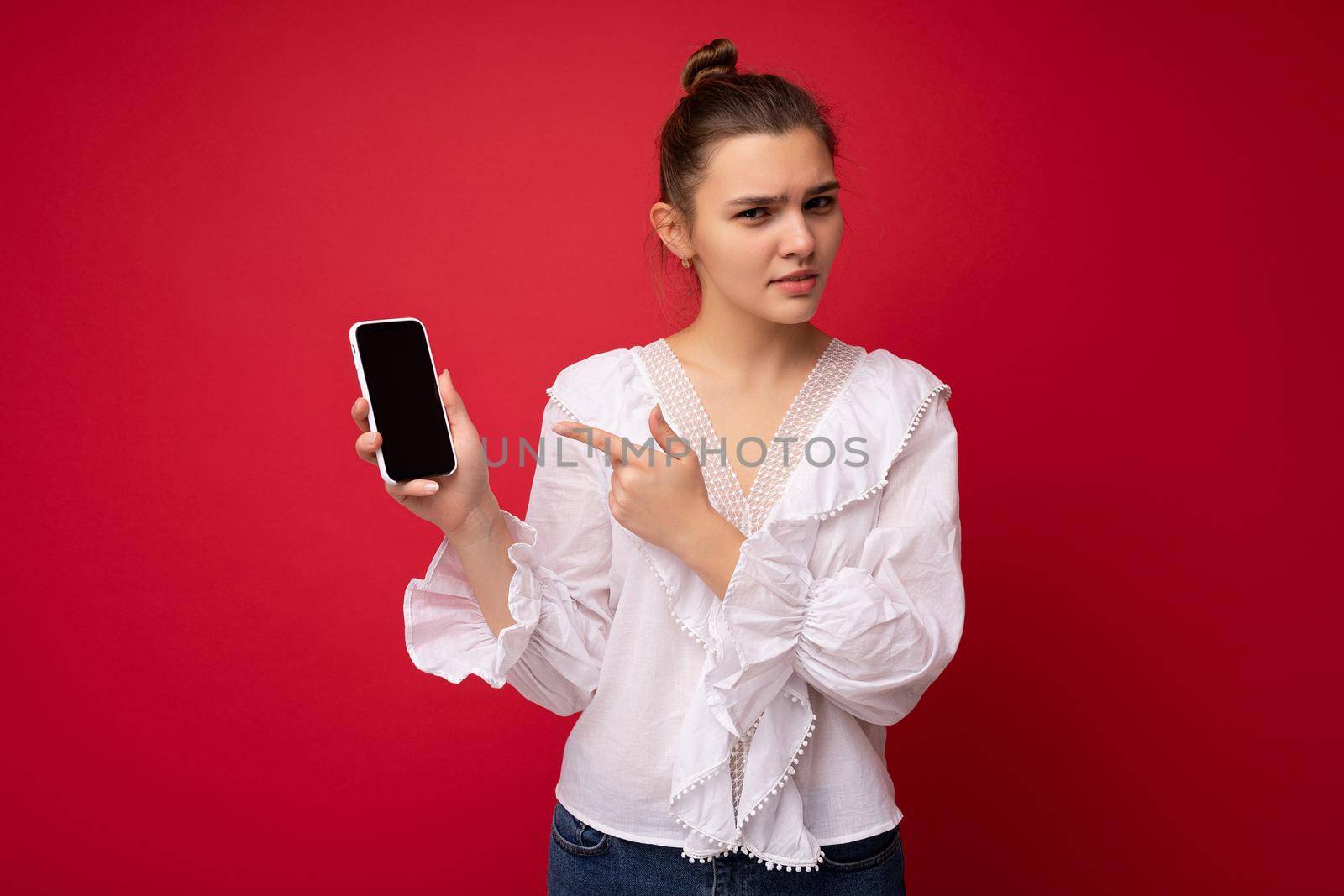 Photo of beautiful upset young woman good looking wearing casual stylish outfit standing isolated on background with copy space holding smartphone showing phone in hand with empty screen display for mockup pointing at gadjet looking at camera by TRMK