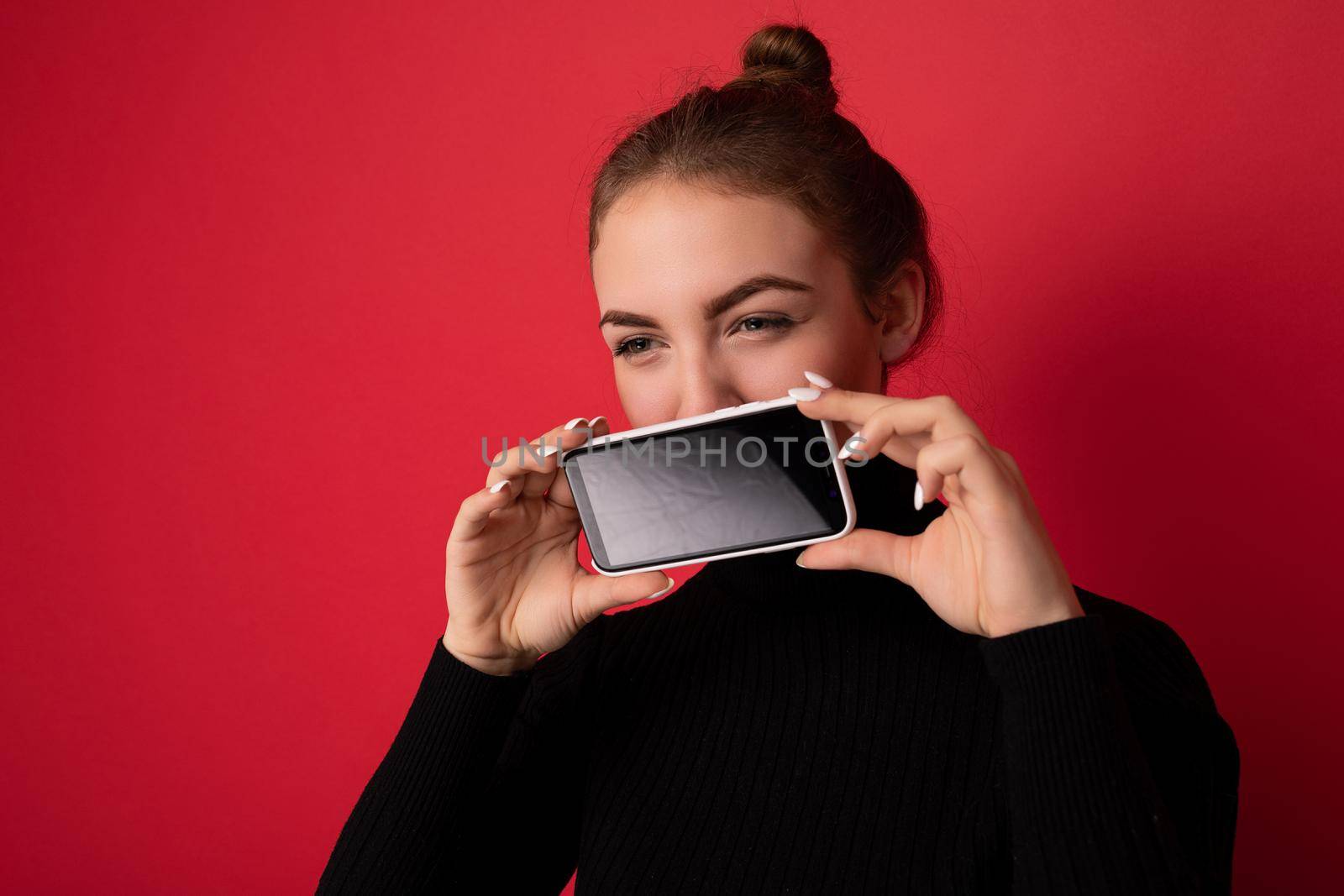 Closeup portrait Photo of beautiful young brunette woman wearing black sweater standing isolated over red background showing mobile phone with empty screen for mockup looking to the side.