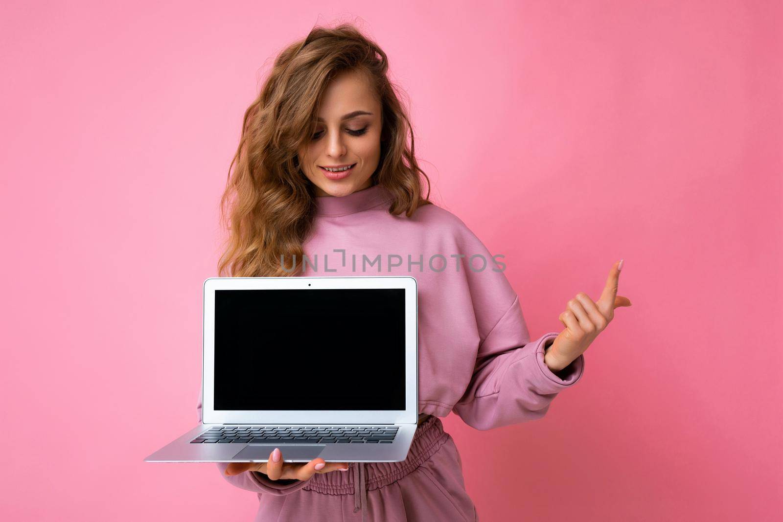 Photo of beautiful blond curly young woman lookind at netbook keyboard holding computer laptop with empty monitor screen with mock up and copy space pointing one finger wearing pink hoodie isolated over pink wall background by TRMK