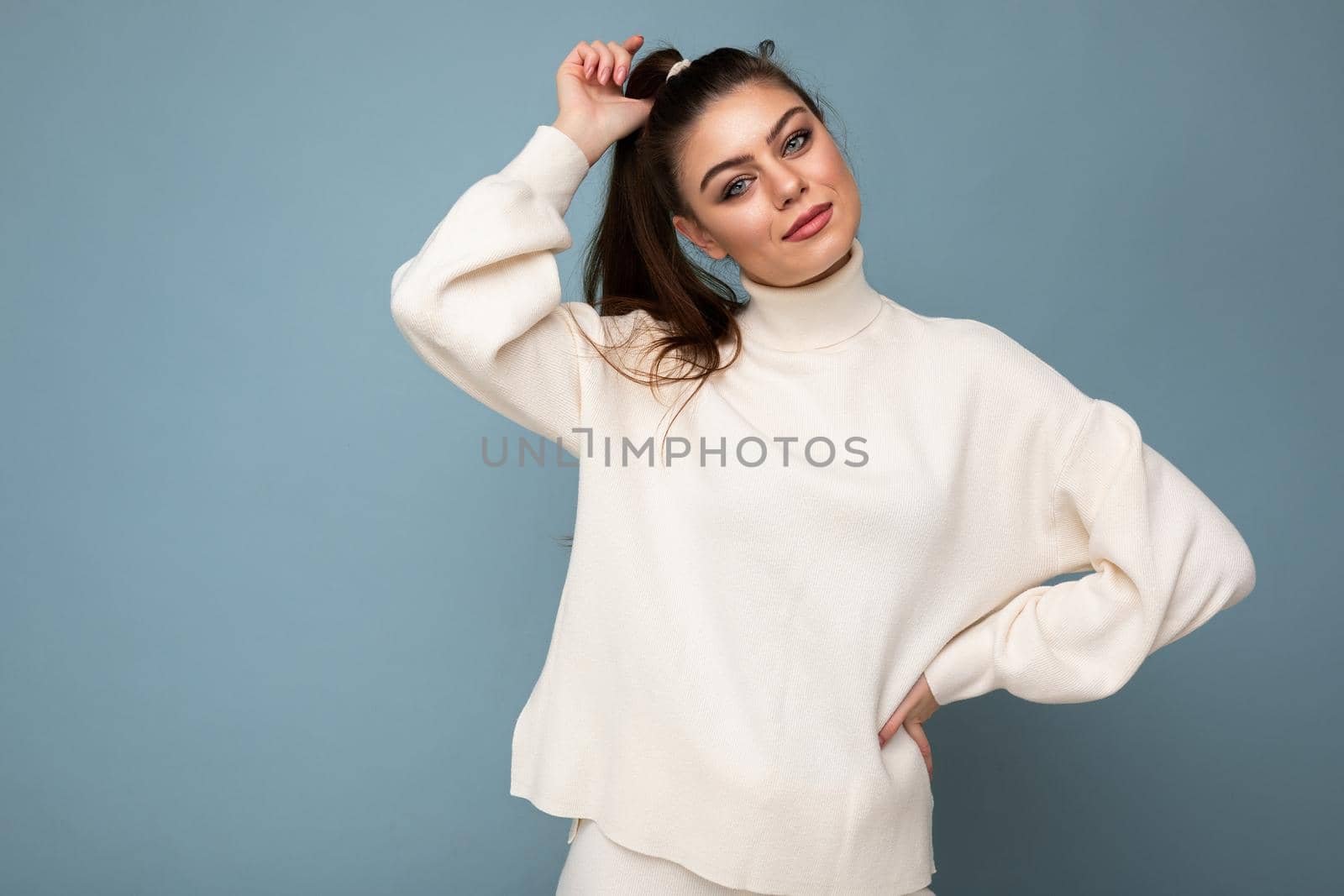 Young pretty european fashion brunette woman wearing casual white sweater isolated on blue background with sincere positive emotional expression on face by TRMK