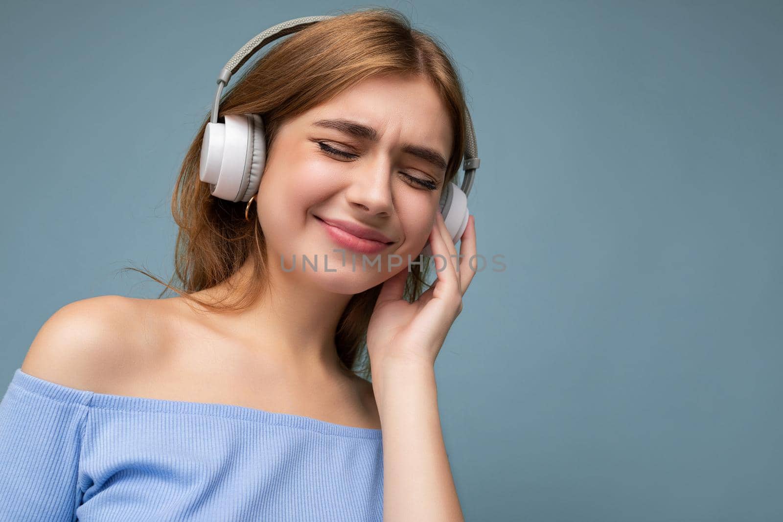 Closeup portrait photo of beautiful positive smiling young blonde woman wearing blue crop top isolated over blue background wall wearing white wireless bluetooth headphones listening to good music and enjoying by TRMK