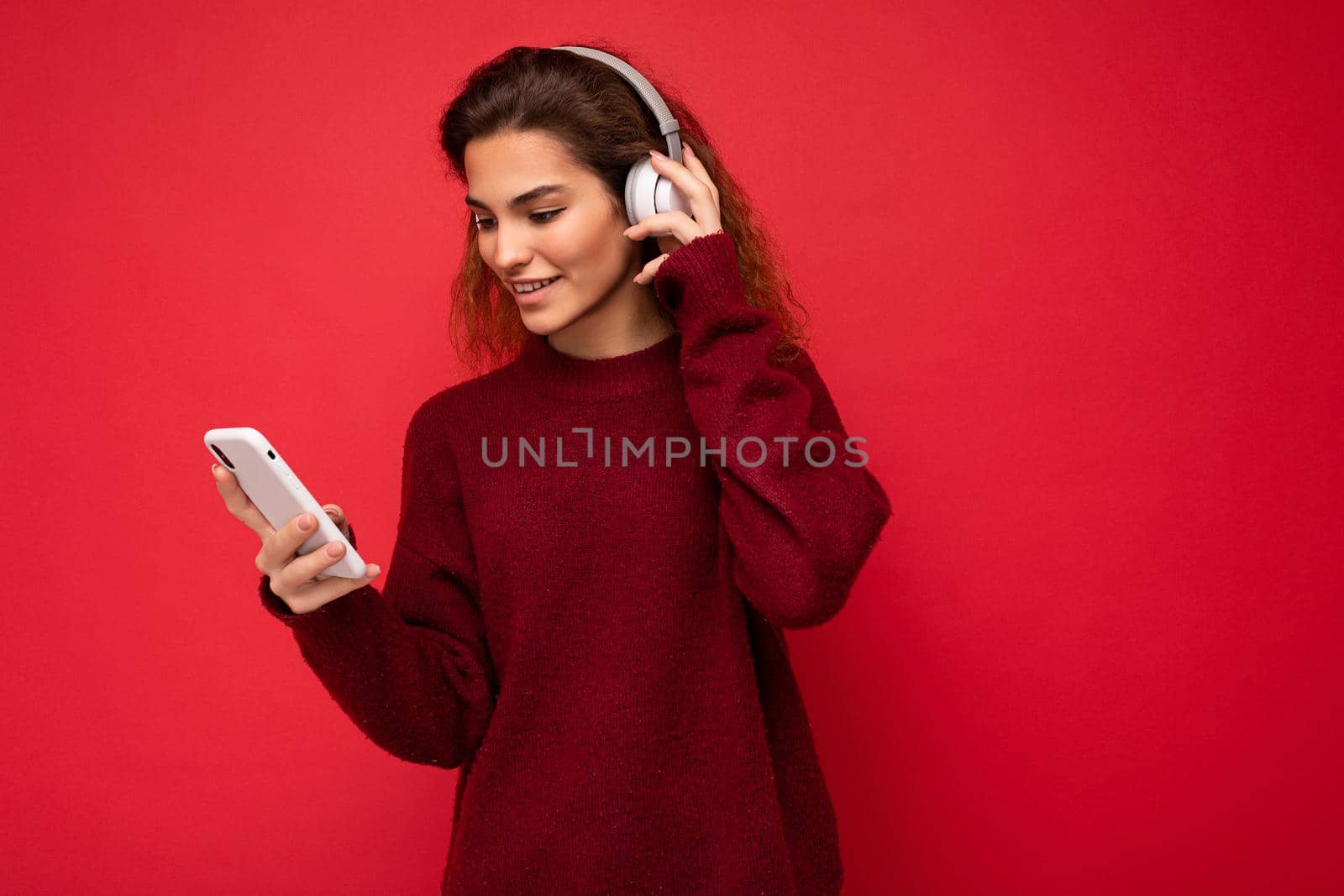 Shot of beautiful young brunette curly woman wearing dark red sweater isolated over red background wall wearing white wireless headphones and listening to music and using mobile phone surfing on the internet looking at smartphone display by TRMK