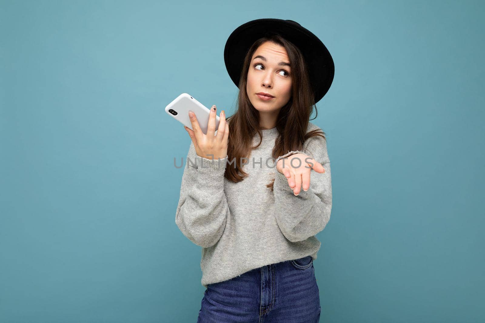 young asking dissatisfied brunette woman wearing black hat and grey sweater holding smartphone looking to the side isolated on background by TRMK