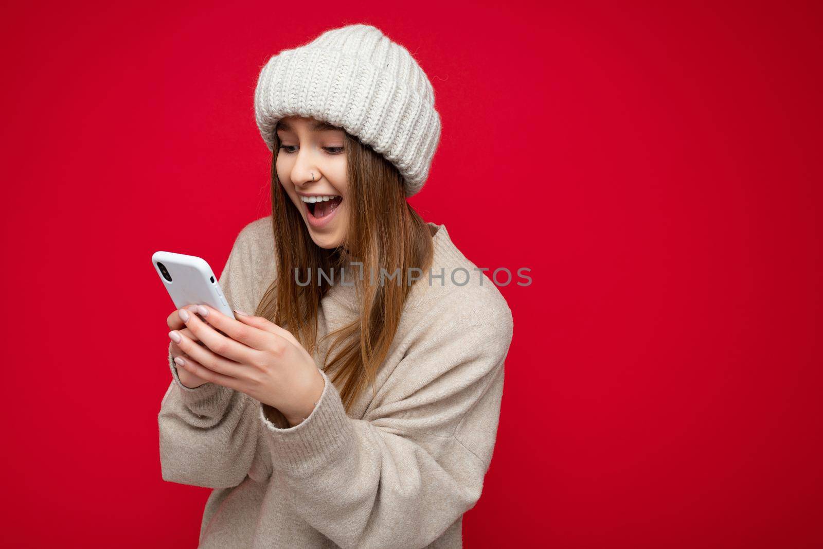 Beautiful amazed young blonde woman wearing casual beige sweater and beige hat isolated over red background holding in hand and using mobile phone communicating online on the internet looking at gadjet display by TRMK