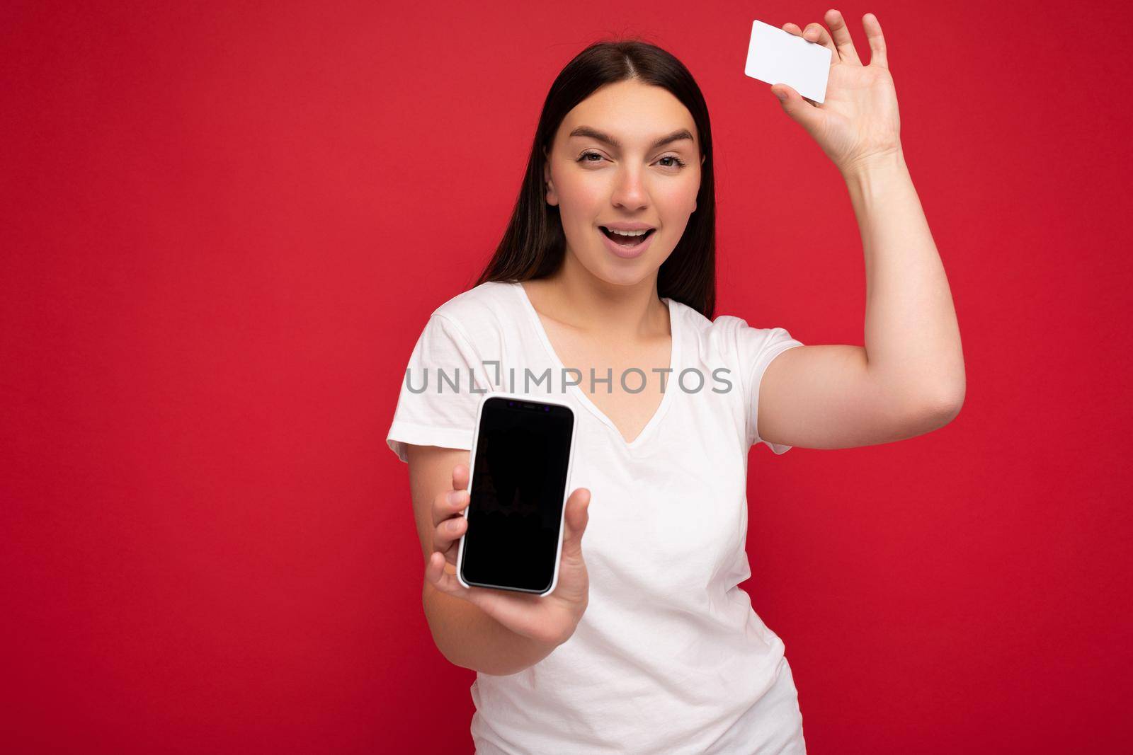 Photo of sexy attractive happy young brunette woman wearing casual white t-shirt isolated over red background with empty space holding in hand mobile phone and showing smartphone with empty screen for mockup and credit card looking at camera.
