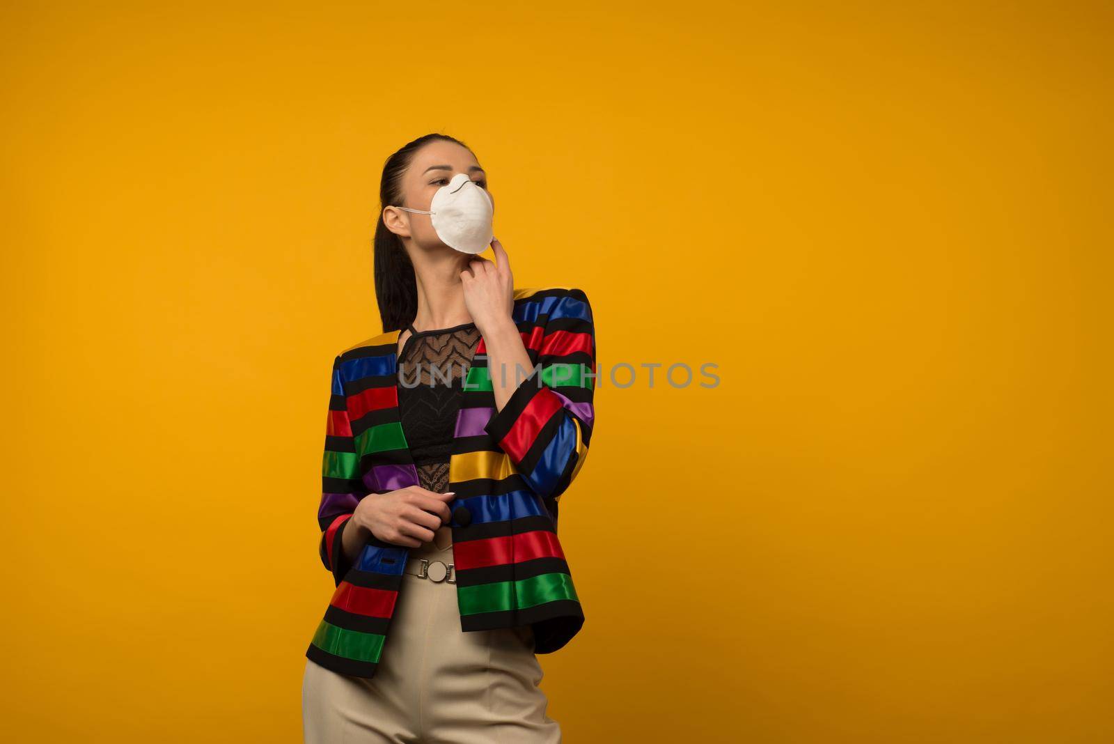 Beautiful slim girl fashion model posing in a protective respirator on a yellow background. LGBT community rainbow color jacket