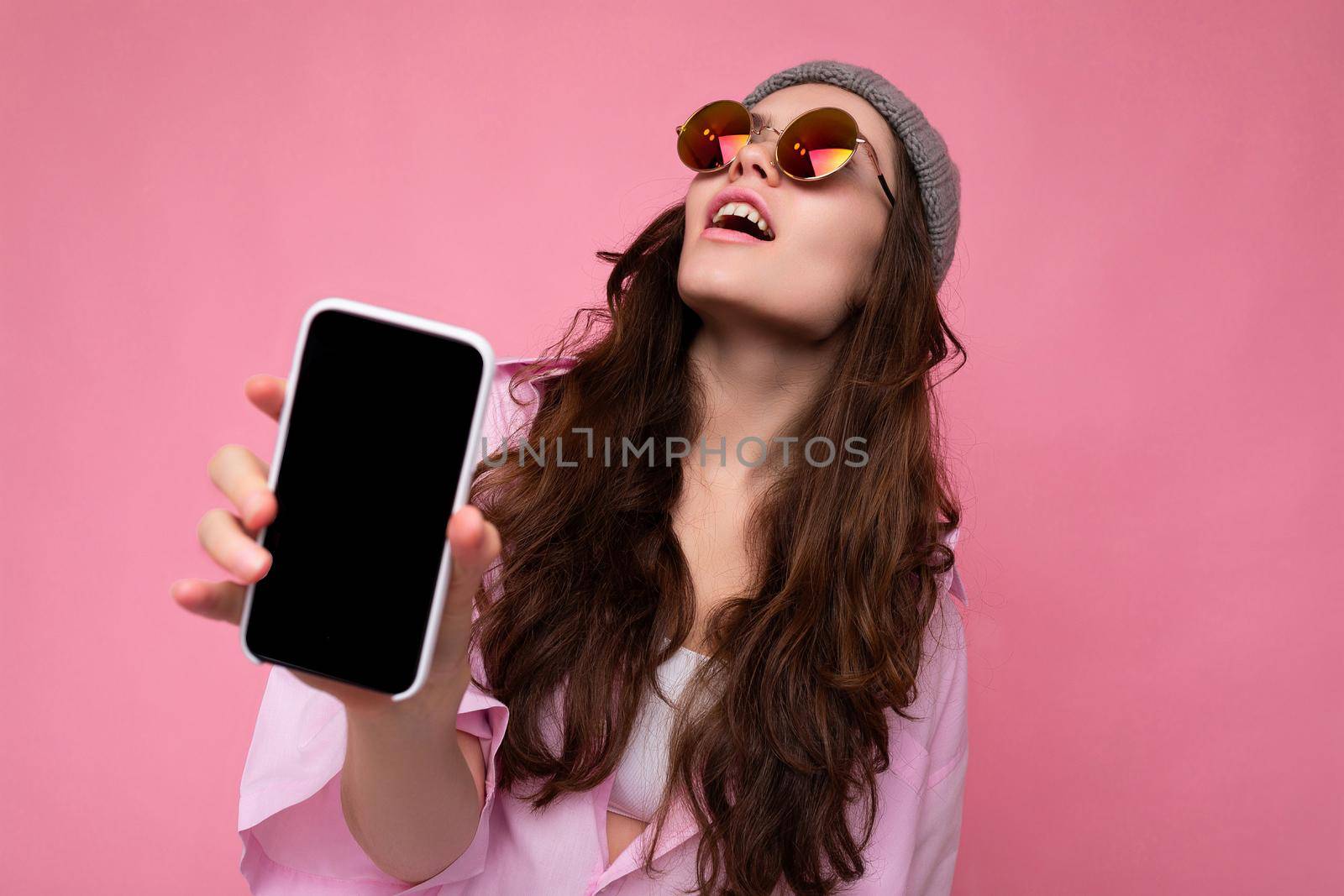 Positive attractive young brunette woman wearing stylish pink shirt grey hat and colourful sunglasses isolated over pink background holding in hand and showing mobile phone with empty display for mockup looking up.