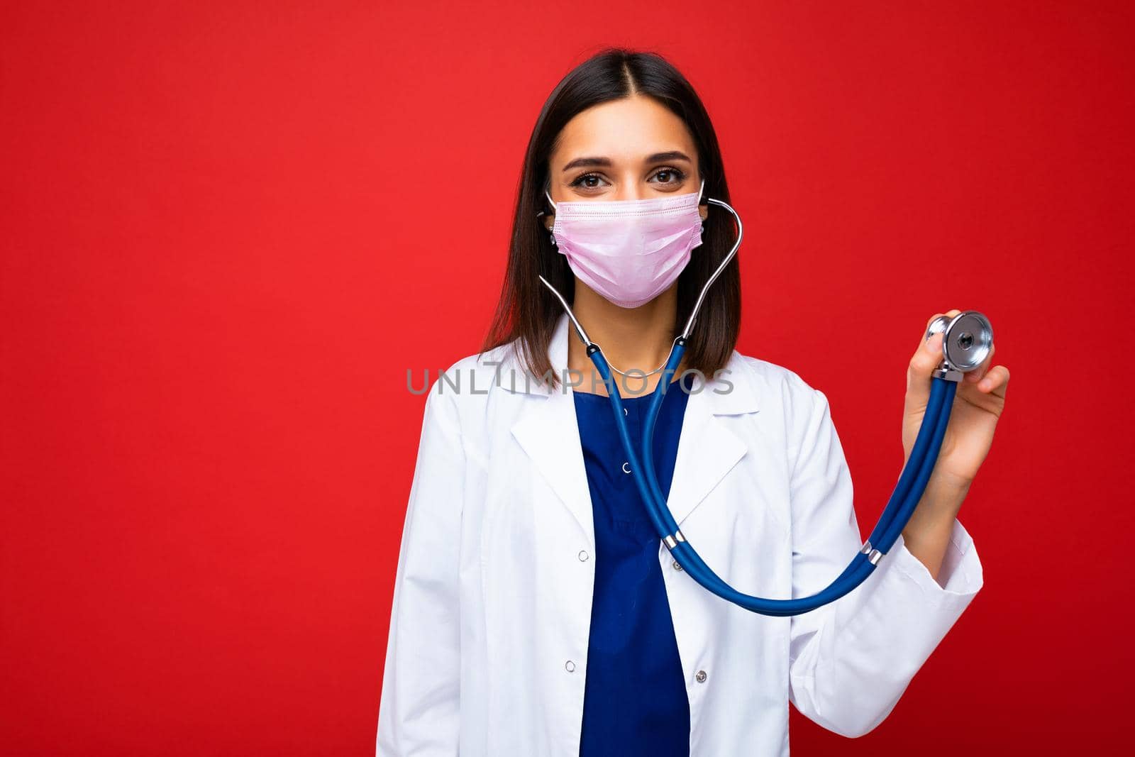 Photo of professional confident young european female doctor in medical mask and white coat, stethoscope over neck isolated on red background by TRMK