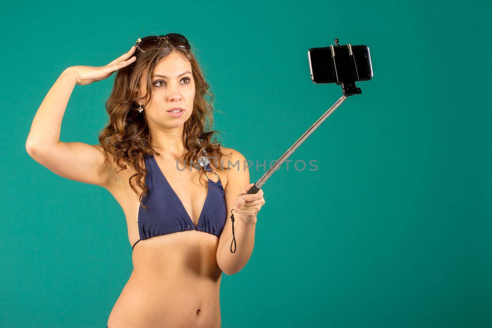 Happy young woman in blue bikini swimsuit taking selfie with smatphone on green background