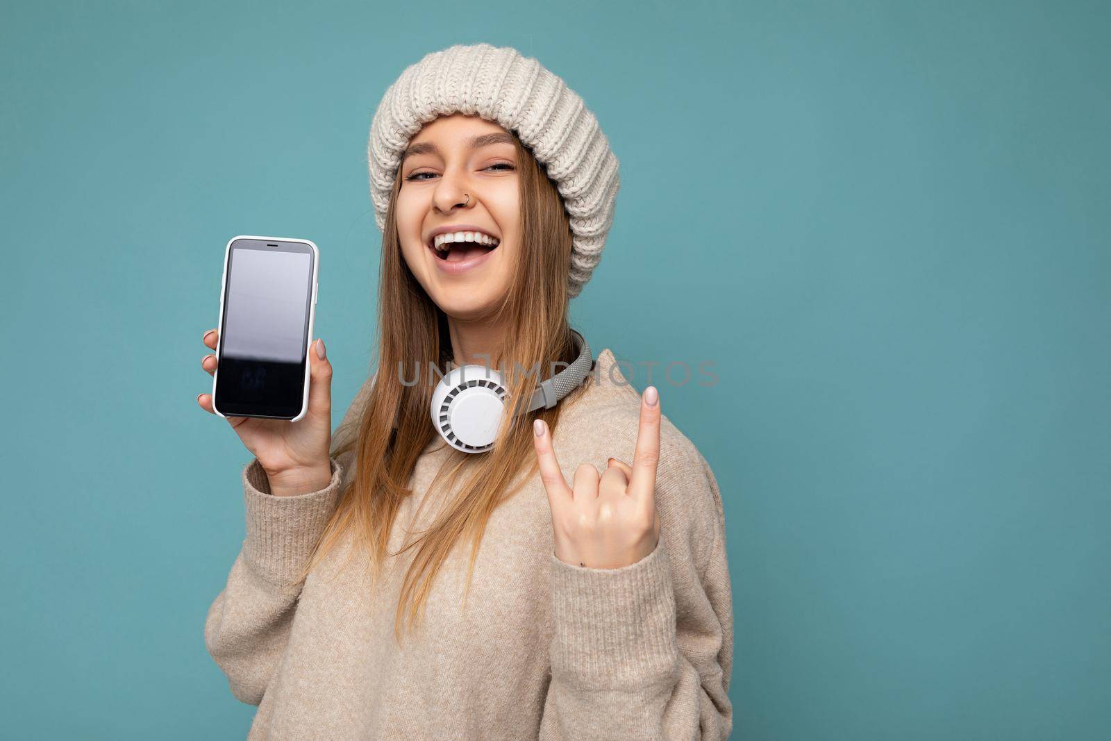 Attractive positive smiling young woman wearing stylish casual outfit isolated on colourful background wall holding and showing mobile phone with empty screen for cutout wearing white bluetooth headphones and having fun looking at camera and showing rock and roll gesture sign by TRMK