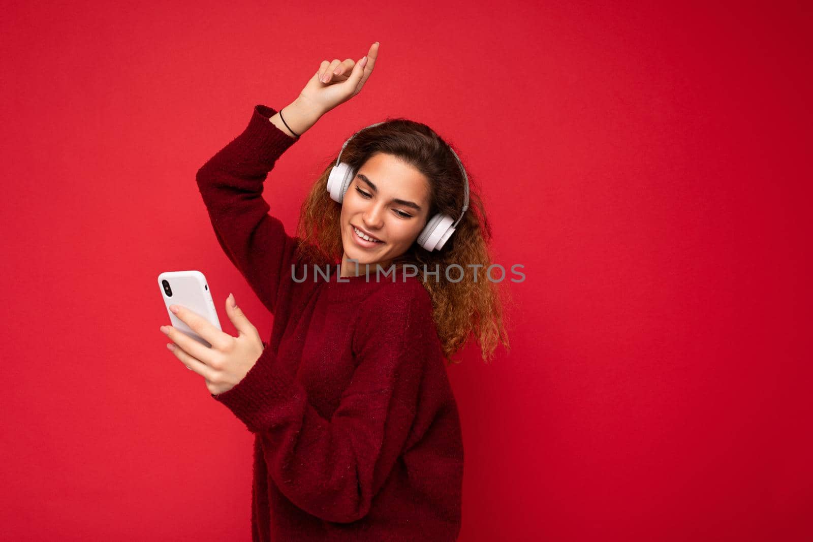Shot of beautiful smiling young brunette curly woman wearing dark red sweater isolated over red background wall wearing white wireless headphones and listening to music and using mobile phone surfing on the internet looking down and dancing. empty space
