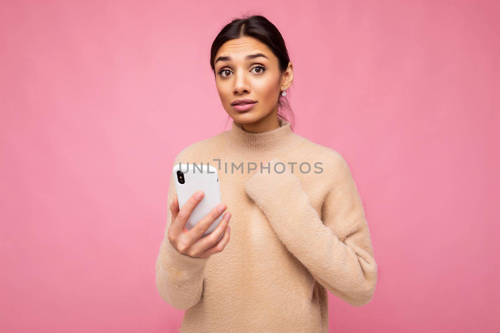 Attractive cute concerned young brunet woman wearing beige warm sweater standing isolated over pink background surfing on the internet via phone looking at camera and keeping hand on heart by TRMK