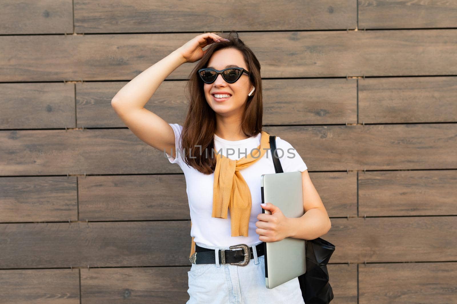 Beautiful smiling charming young brunet woman looking at camera holding computer laptop and sunglasses in white t-shirt and light blue jeans in the street by TRMK