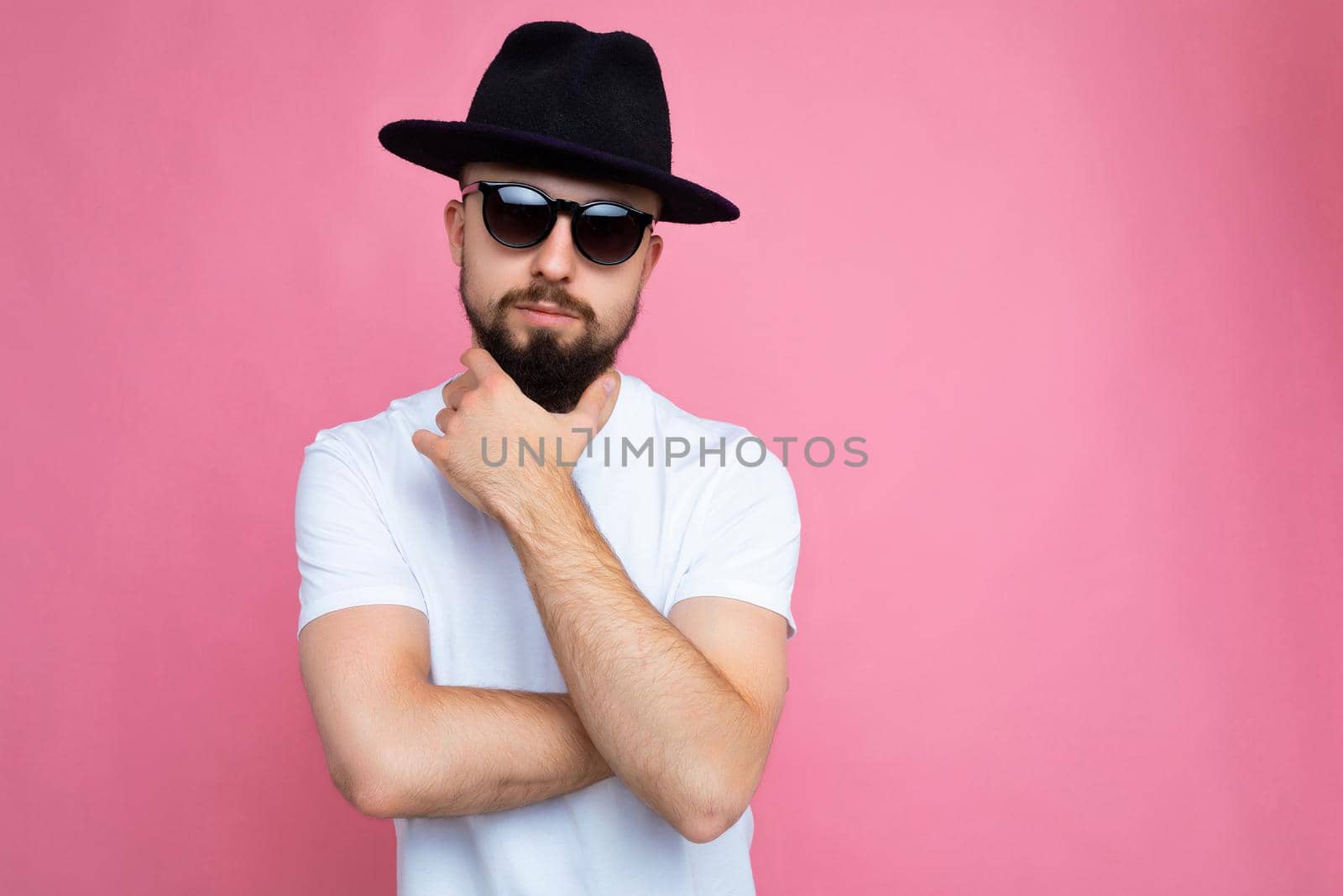 Photo shot of sexy handsome brunette unshaven young man with beard wearing casual white t-shirt for mockup black hat and stylish sunglasses isolated over pink background wall looking at camera. empty space
