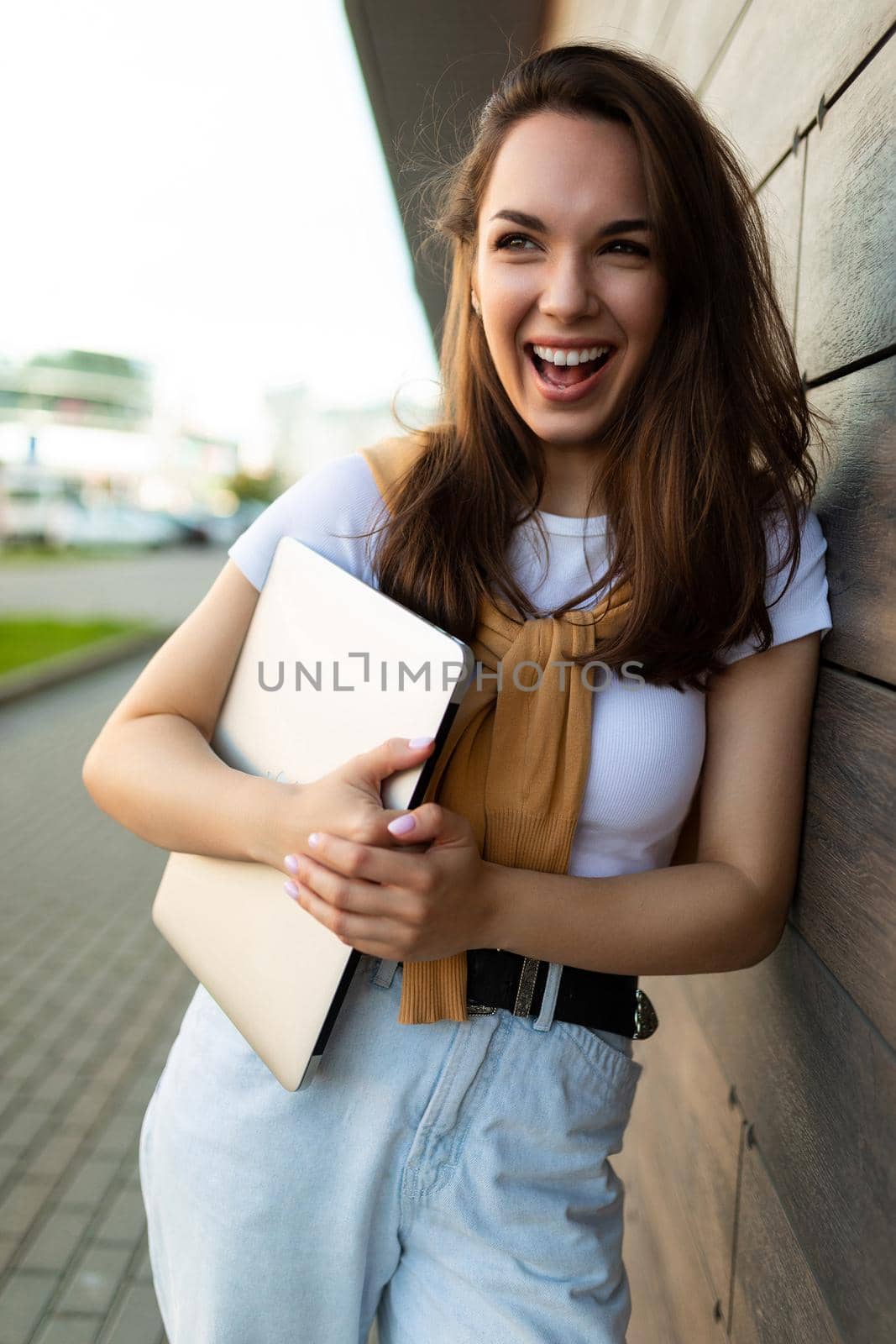 beautiful happy brunet young woman looking to side in the street with laptop computer in hands with white t-shirt and blue jeans by TRMK