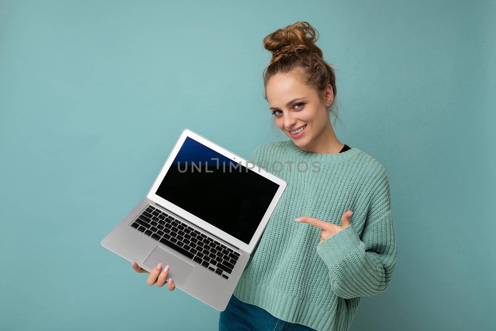 beautiful dark blond young woman with gathered curly hair looking at camera holding computer laptop with empty monitor screen with mock up and copy space wearing blue longsleeve isolated on light blue wall background.