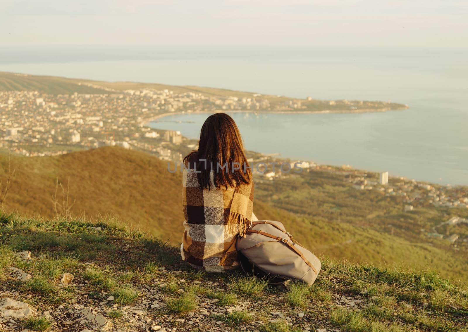 Young woman wrapped in plaid sitting on peak of mountain and enjoying view of sea