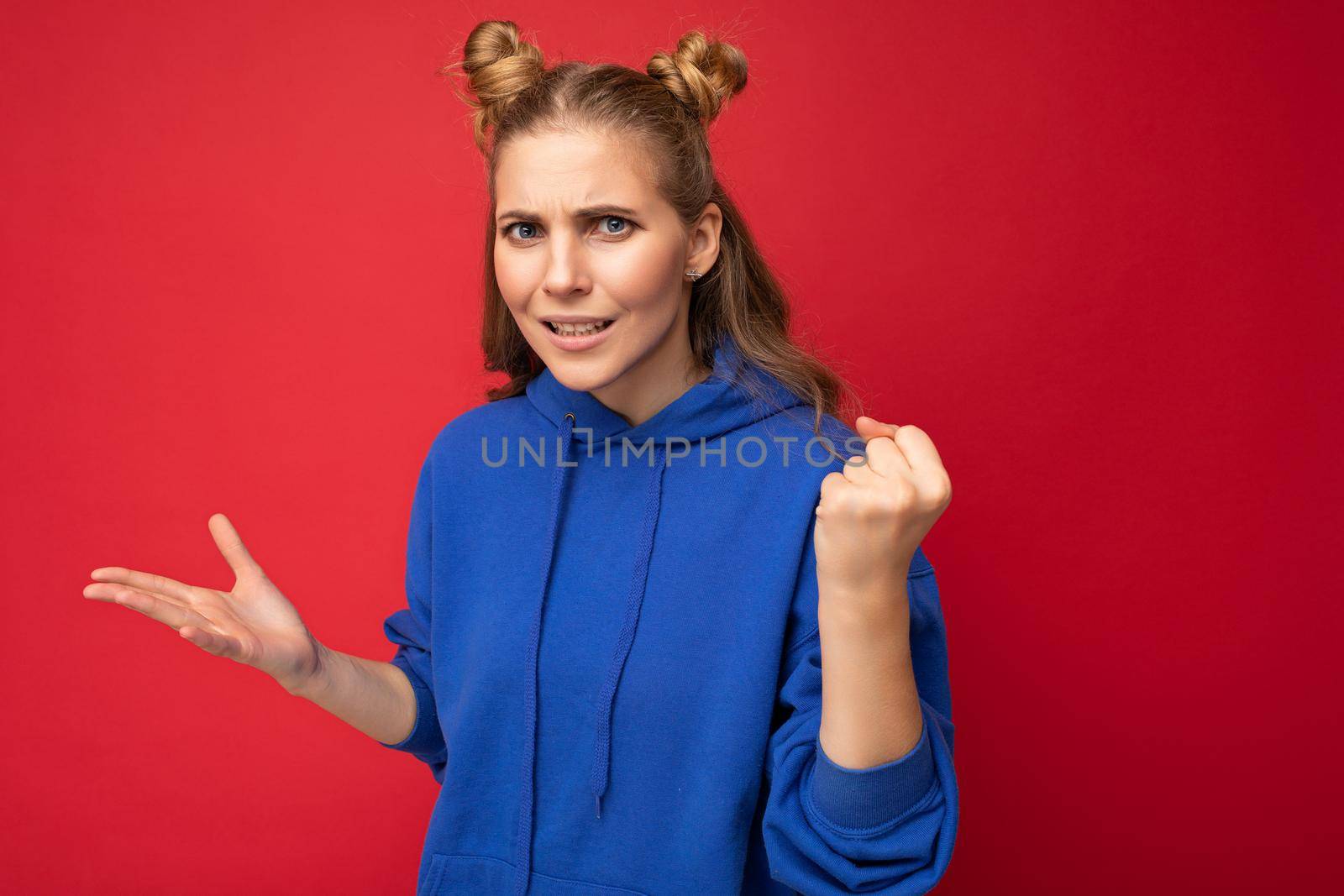 Photo of emotional agressive young beautiful blonde woman with two horns with sincere emotions wearing hipster bright blue hoodie isolated over red background with free space and showing fist by TRMK