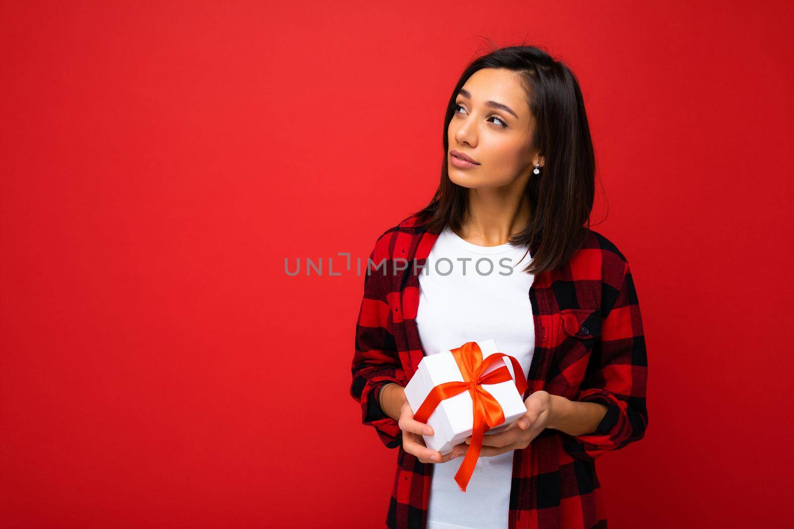 Photo shot of pretty positive young brunette woman isolated over red background wall wearing white casual t-shirt and red and black shirt holding white gift box with red ribbon and looking to the side. Free space