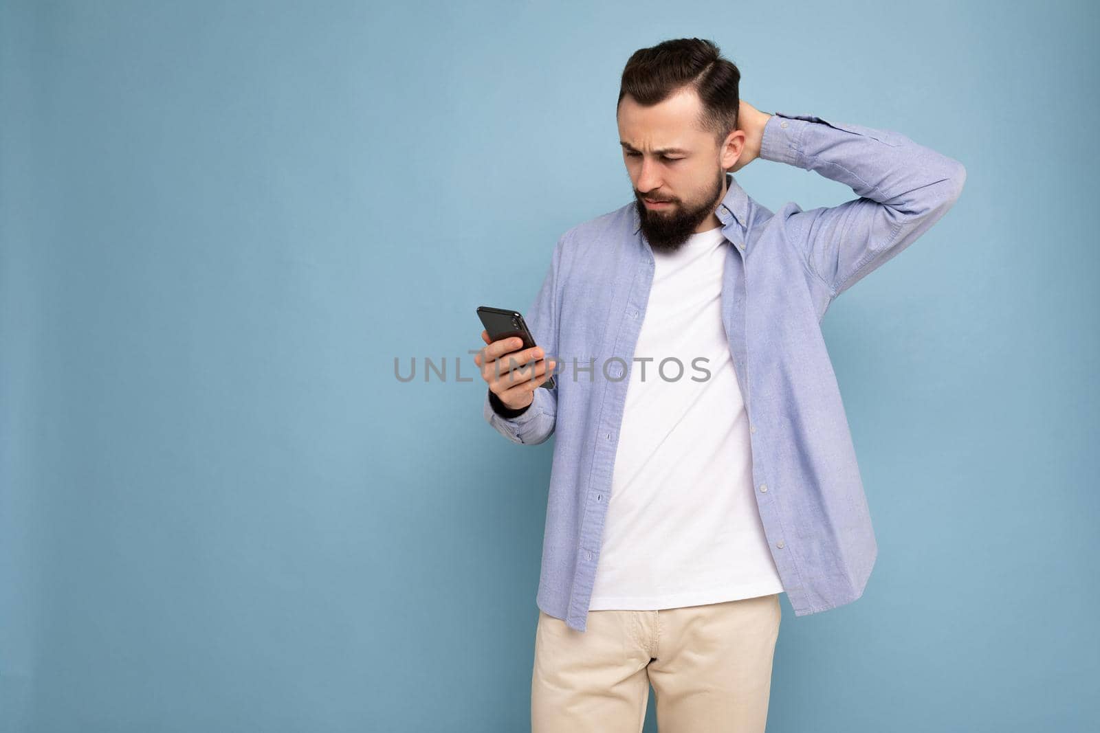 side-profile Photo shot of handsome good looking young man wearing casual stylish outfit poising isolated on background with empty space holding in hand and using mobile phone messaging sms looking at smartphone display screen and thinking over by TRMK