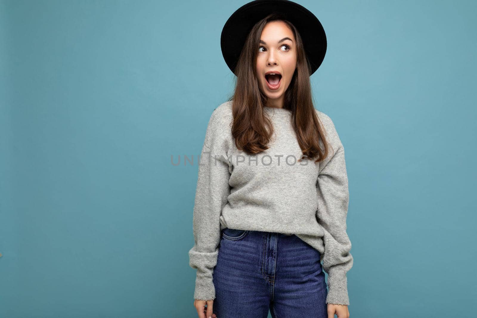 Young beautiful brunet woman. Trendy lady in casual grey sweater and stylish black hat. Positive female shows facial sincere emotions. Funny model isolated on blue background with free space.
