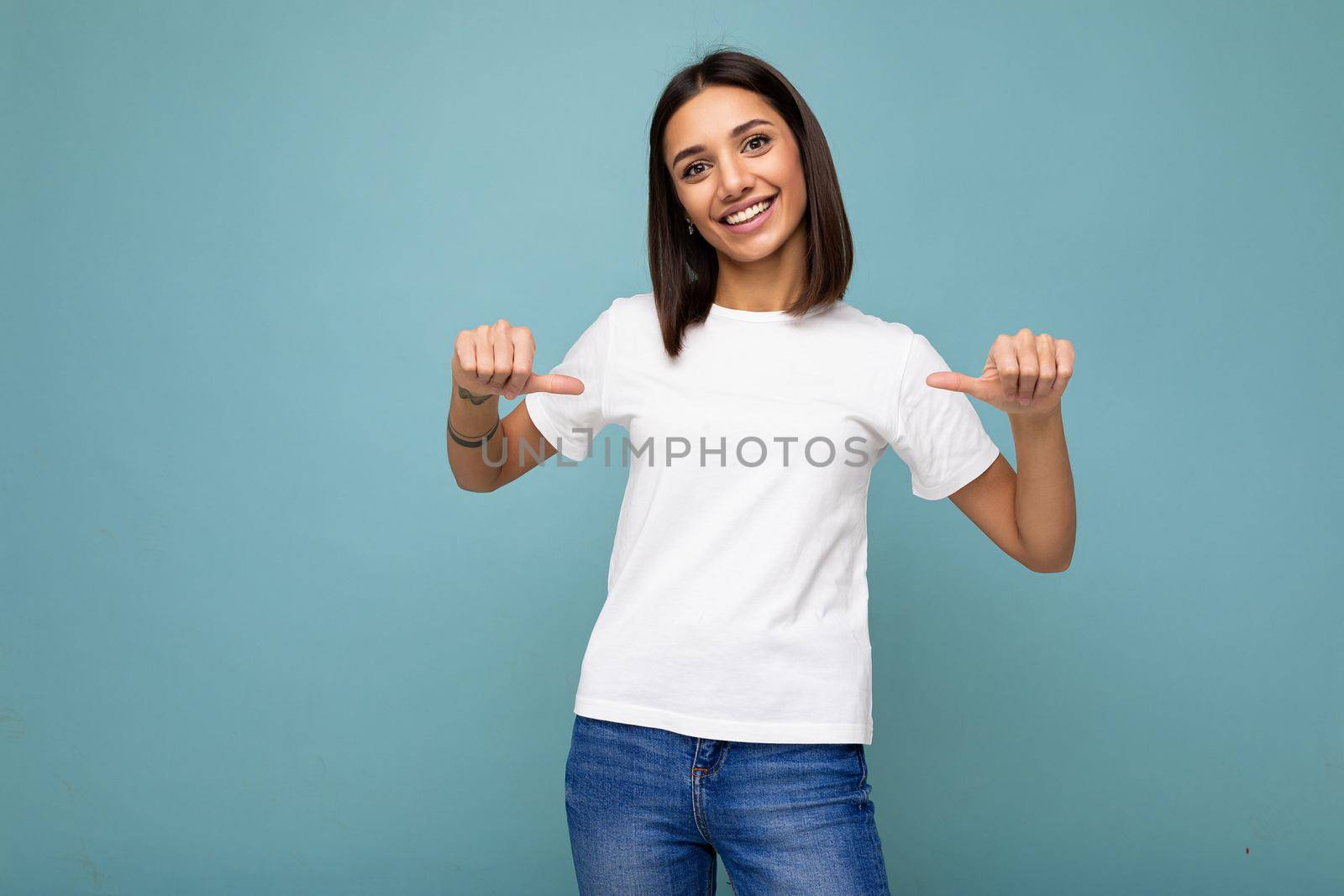 Portrait of positive happy smiling young pretty cute brunette woman with sincere emotions wearing casual white t-shirt for mockup isolated over blue background with copy space and pointing at empty space.