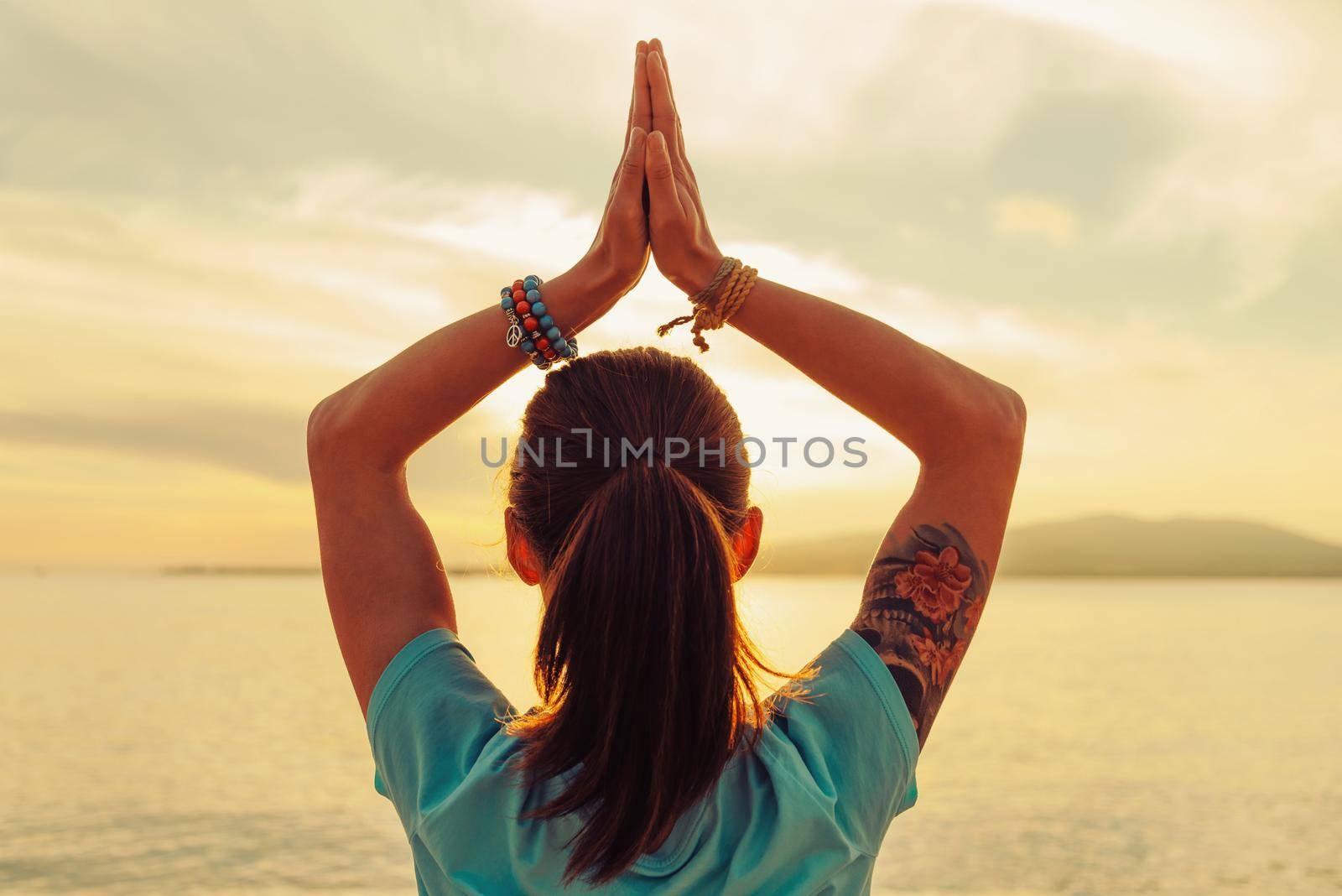 Young woman doing yoga exercise on beach at sunset in summer, rear view