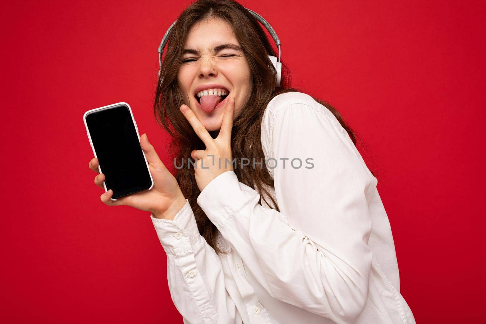 Attractive sexy positive young brunette curly woman wearing white shirt isolated on red background wall holding and showing mobile phone with empty display wearing white bluetooth headphones listening to music showing fingers and tongue.