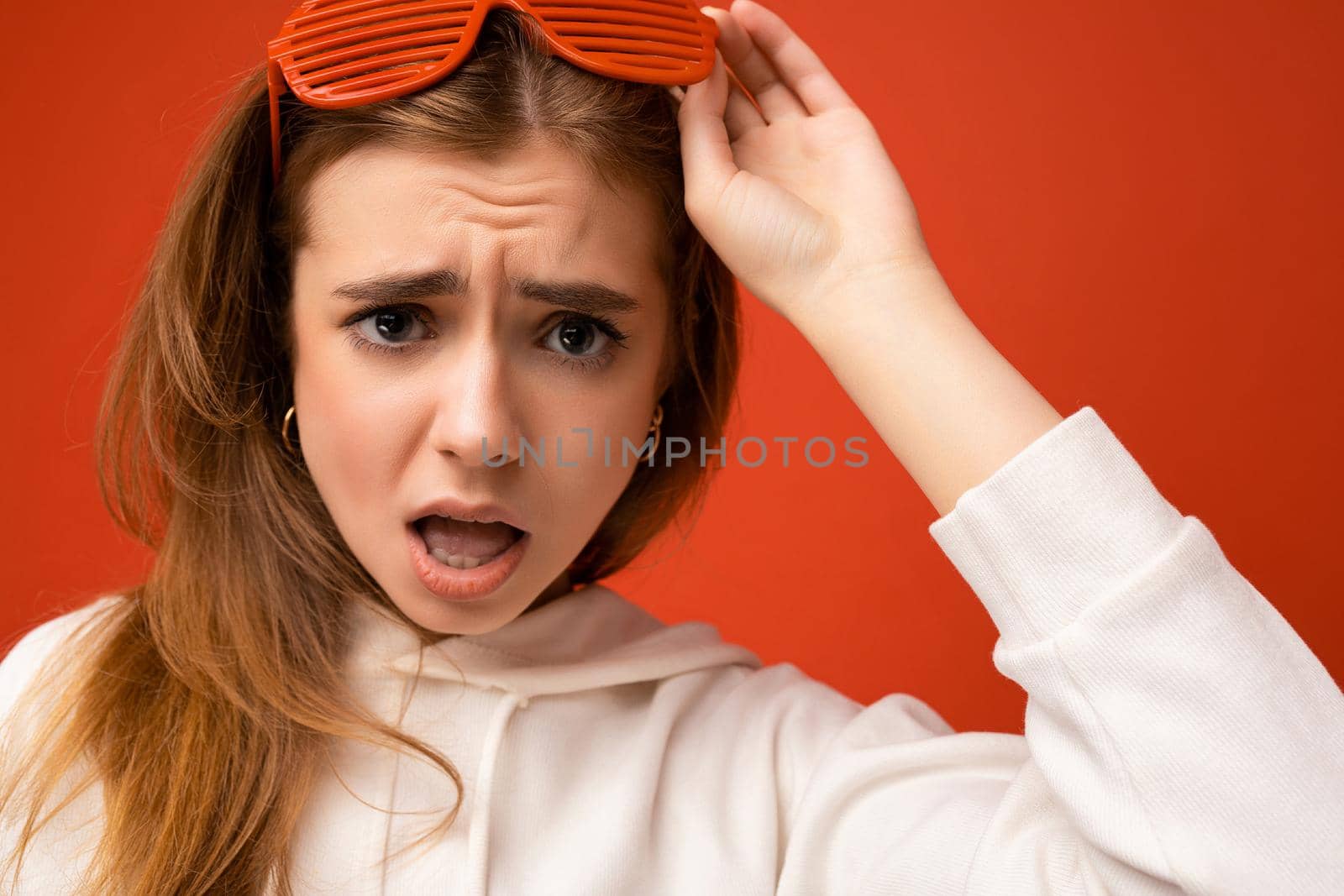 Closeup photo shot of beautiful sad shocked young blonde woman isolated over colourful background wearing casual white hoodie and stylish glasses looking at camera with open mouth by TRMK